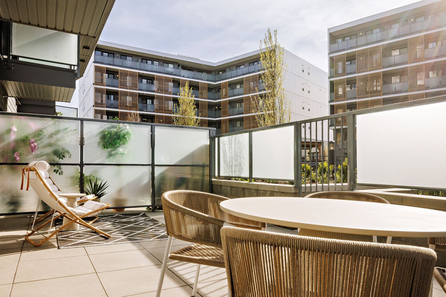 Modern apartment balcony with wooden deck chairs and glass railing overlooking residential buildings.