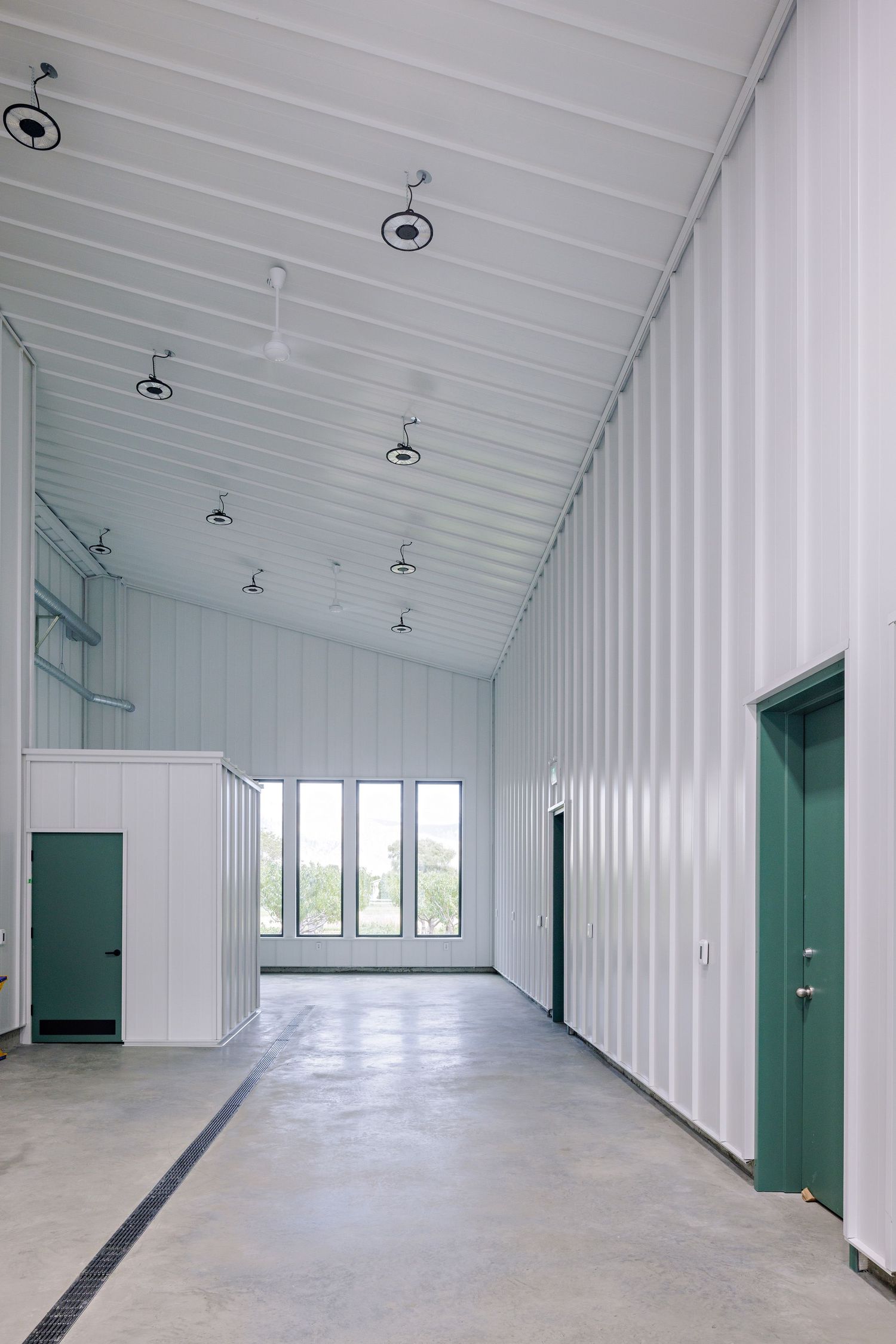 Modern industrial hallway with white corrugated walls, green doors, high ceiling lights, and concrete flooring.