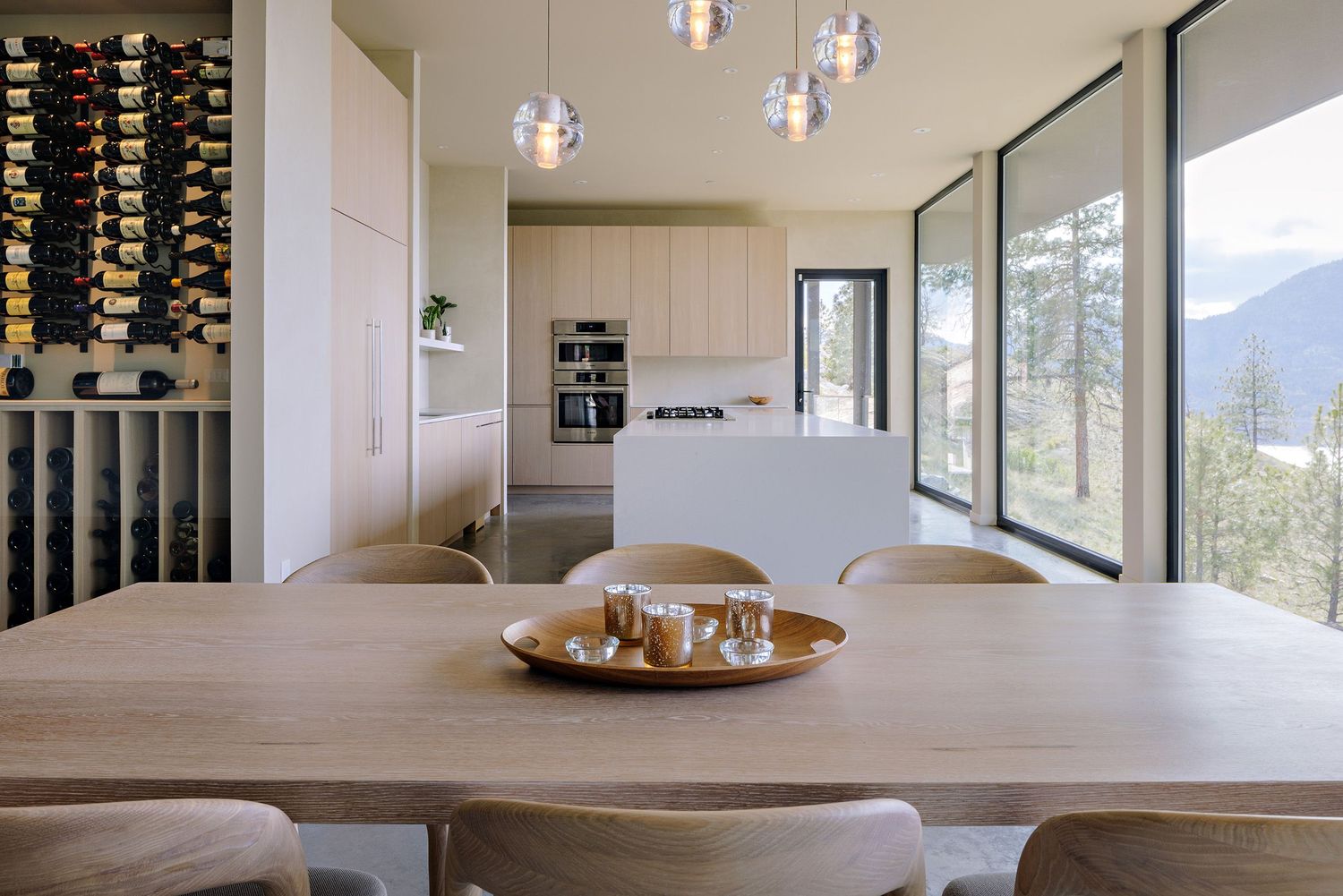Modern dining area with wine storage features wood table, pendant lights and mountain views through large windows.