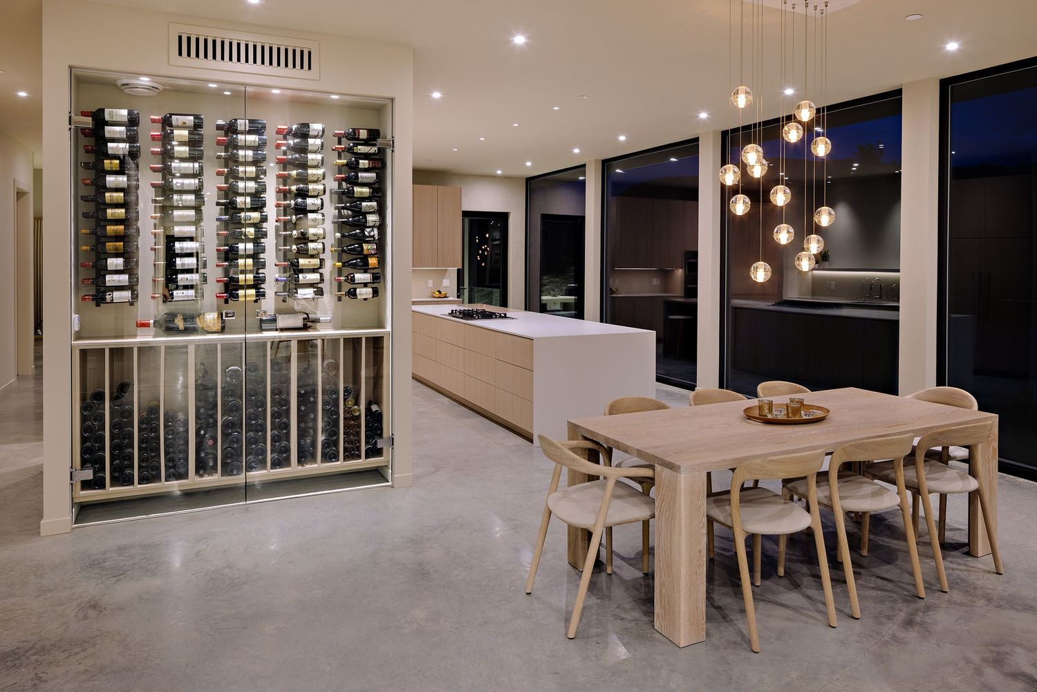 Modern dining room with illuminated glass wine wall, wood table, pendant lights, and polished concrete floors.