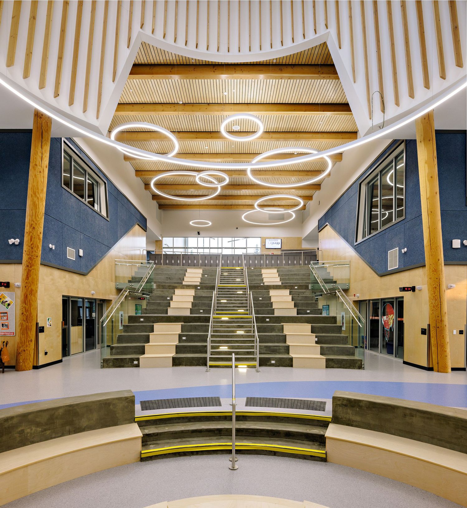Modern auditorium space with tiered seating and distinctive circular lighting fixtures mounted on wooden ceiling panels.