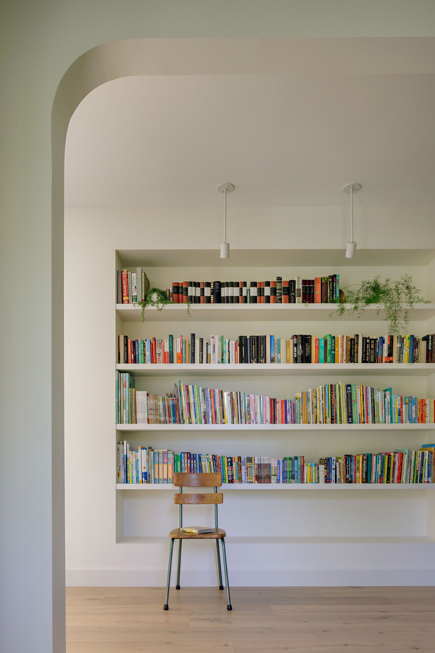 Long white wall-mounted bookshelf filled with colorful books and a single wooden chair in a minimalist room.