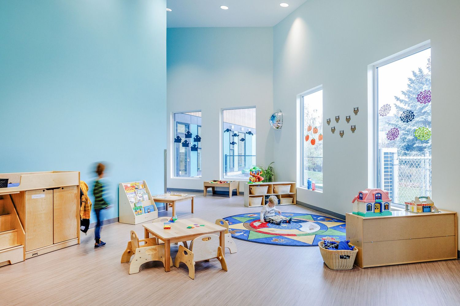 Modern daycare center interior with natural light featuring play areas, wooden furniture and colorful decorations.