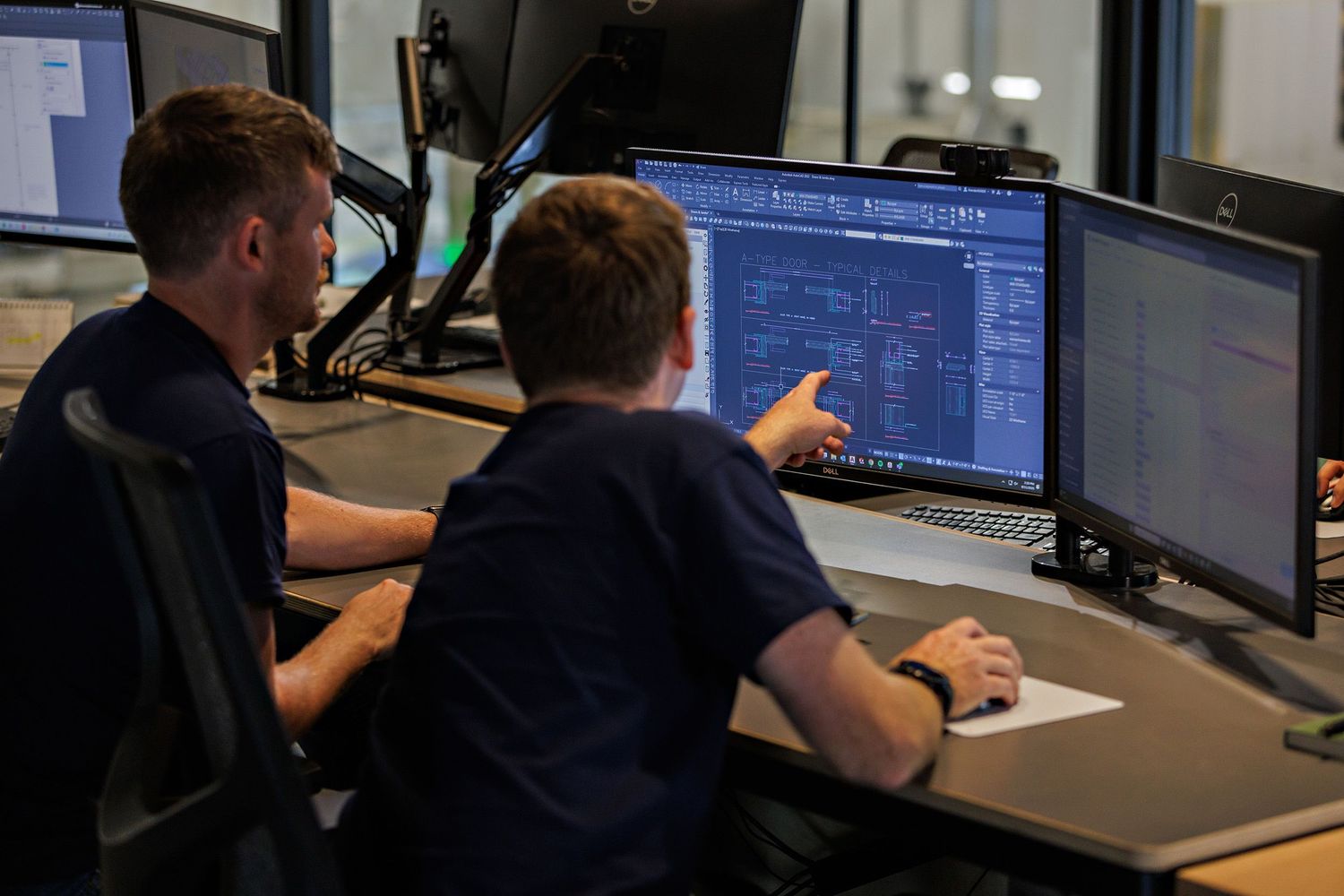 Students work at computer stations in a classroom lab setting with multiple monitors and workstations visible.
