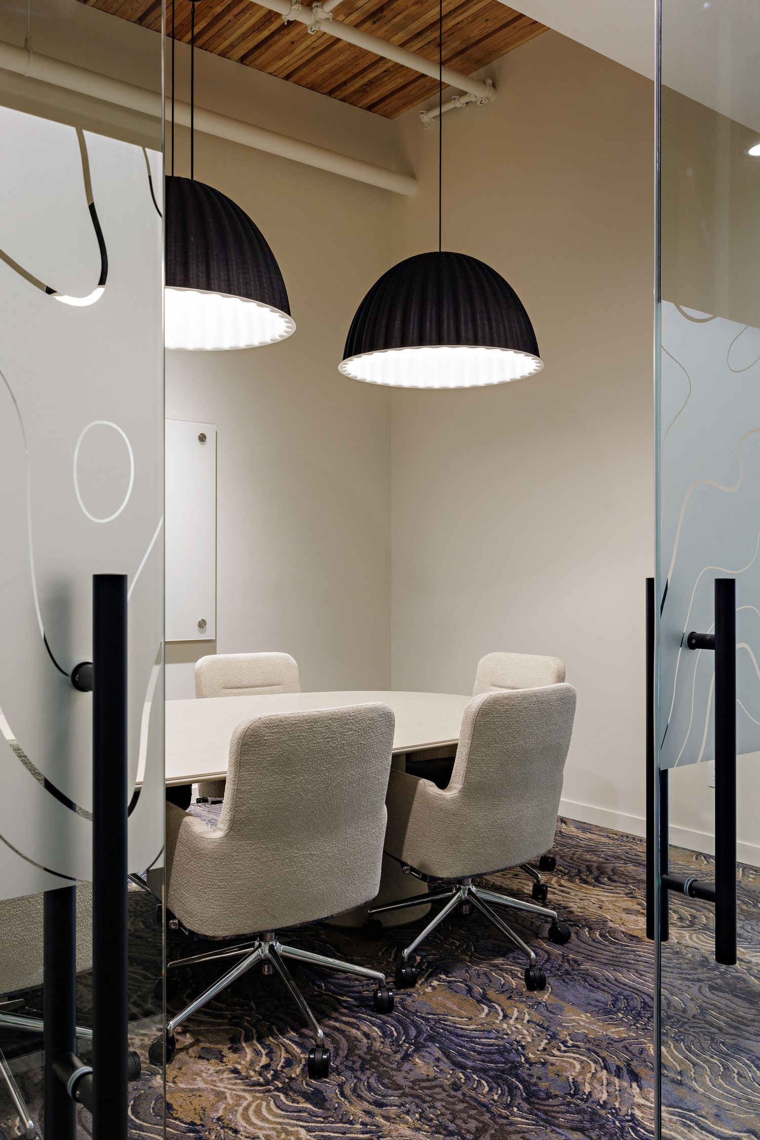 Modern office meeting room with glass walls, two white upholstered chairs, and large black dome pendant lights.