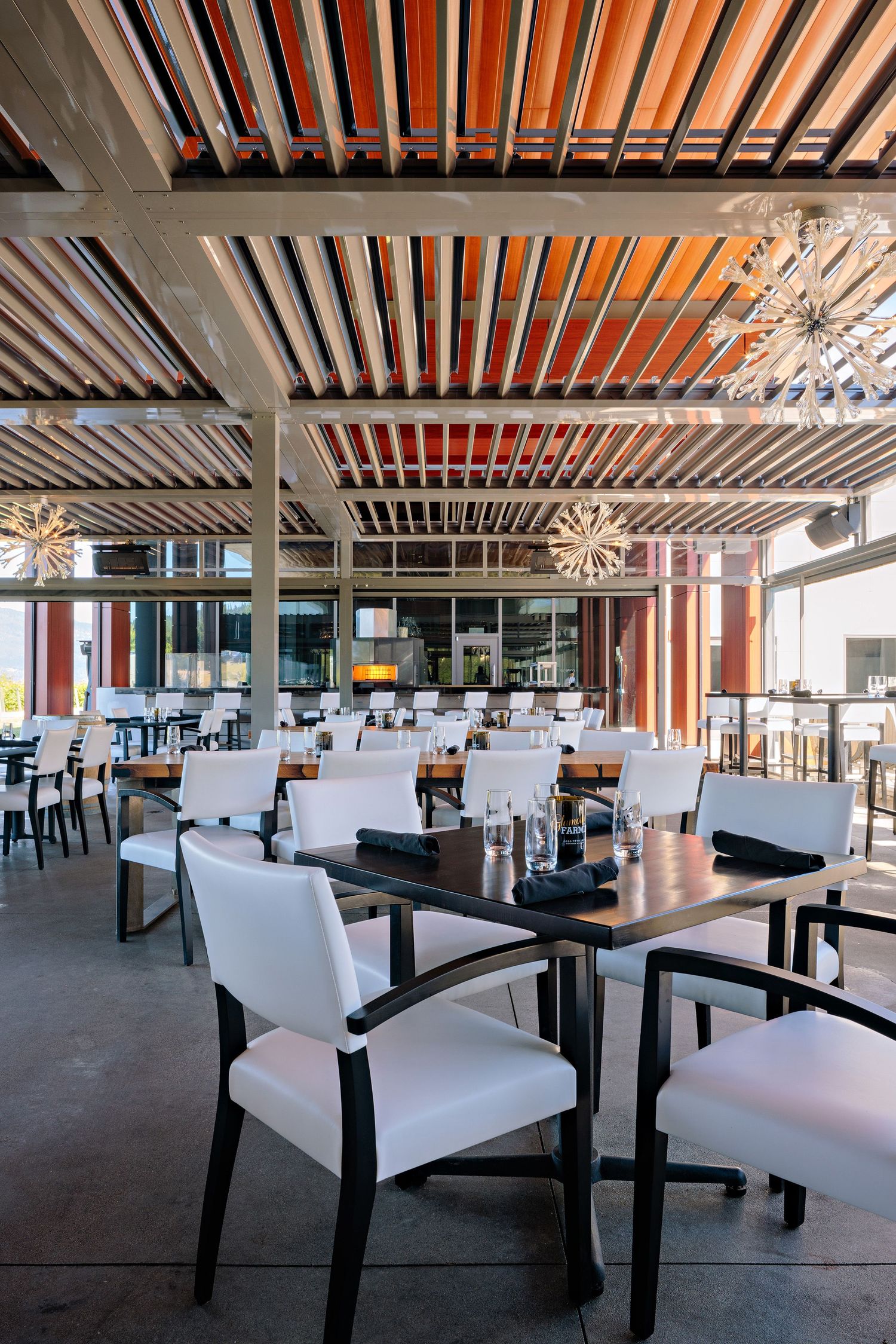 Modern restaurant interior with white chairs, dark tables, and metal ceiling beams illuminated by hanging chandeliers.