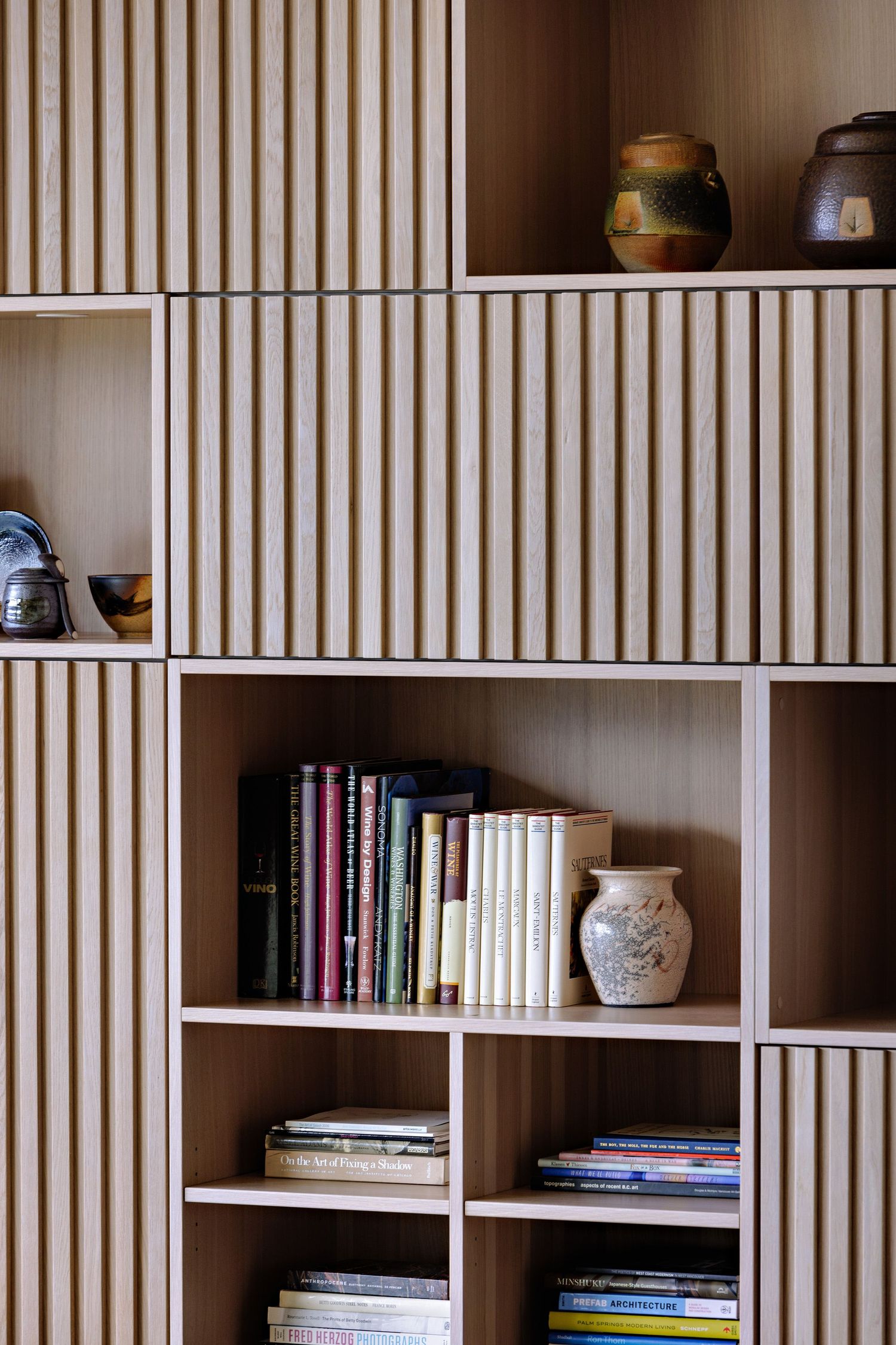 Modern white shelving unit with wooden slat doors displays books and ceramic pottery in a minimalist Scandinavian style.