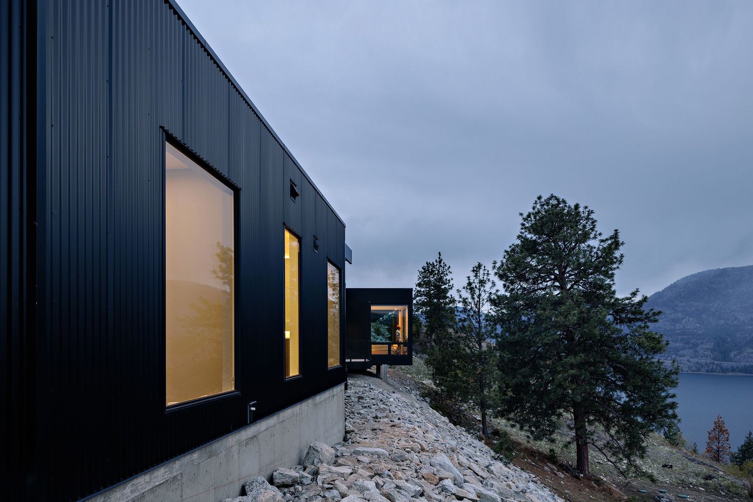 Modern black cabin with illuminated windows perched on rocky mountainside overlooking a serene lake at dusk.