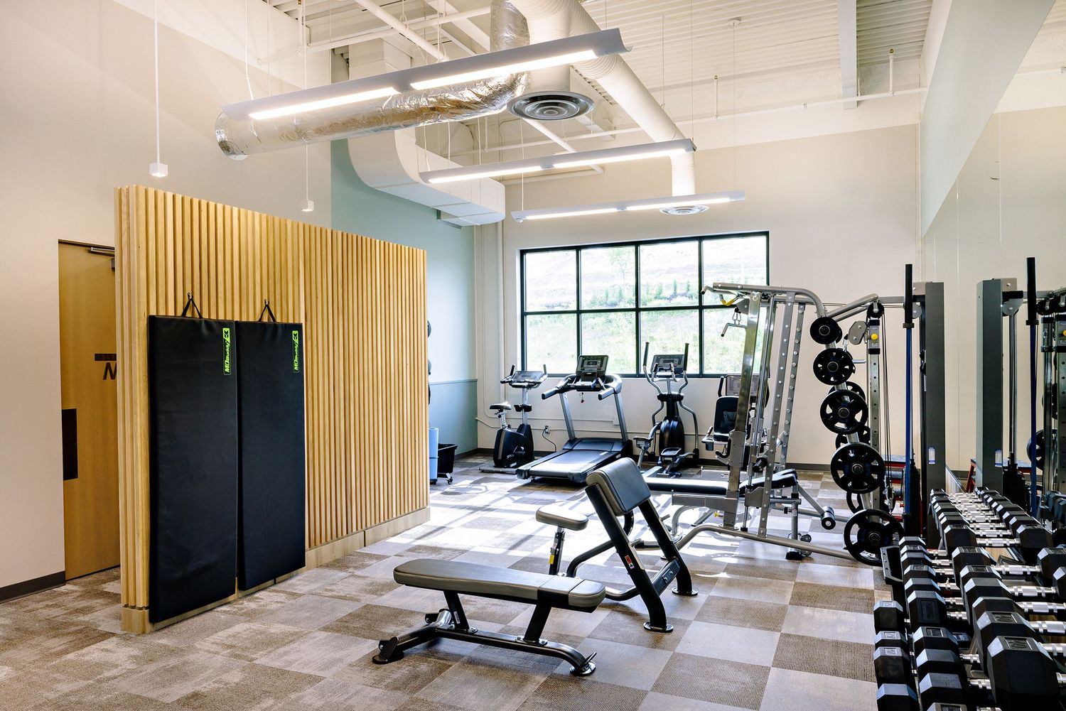 Modern fitness center with weight equipment, black exercise benches, and wooden accent wall featuring checkered flooring.