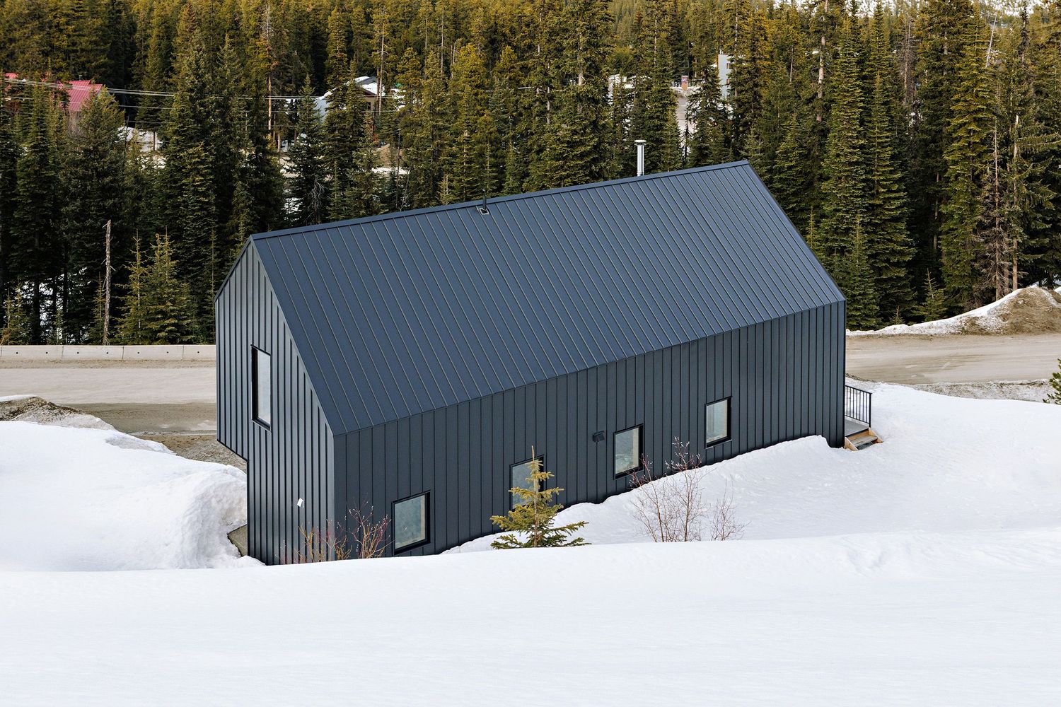 A modern metal barn or storage building with a grey exterior sits in a snowy landscape surrounded by evergreen trees.