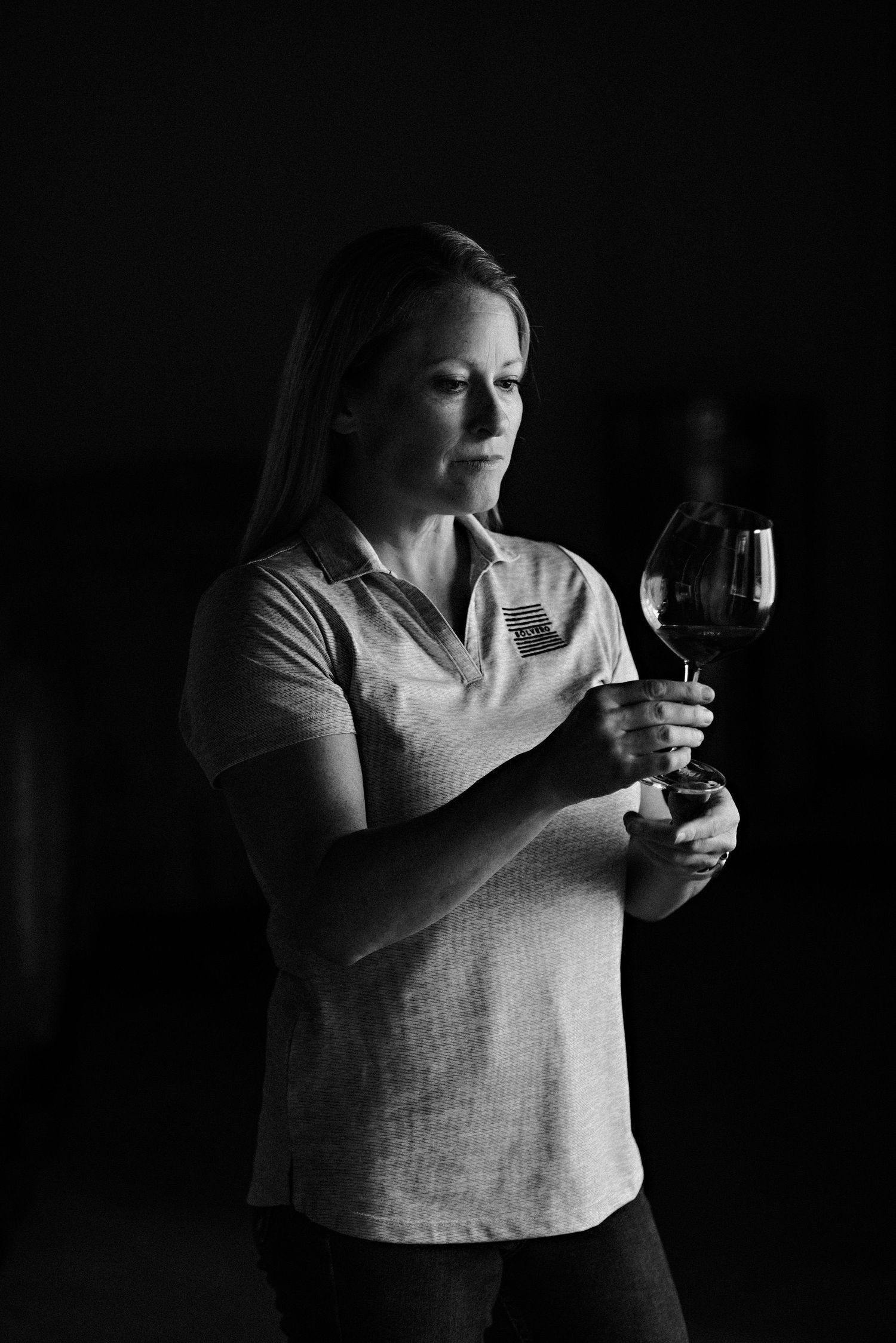 A dramatic black and white portrait of a person examining a glass of red wine in contemplative lighting.