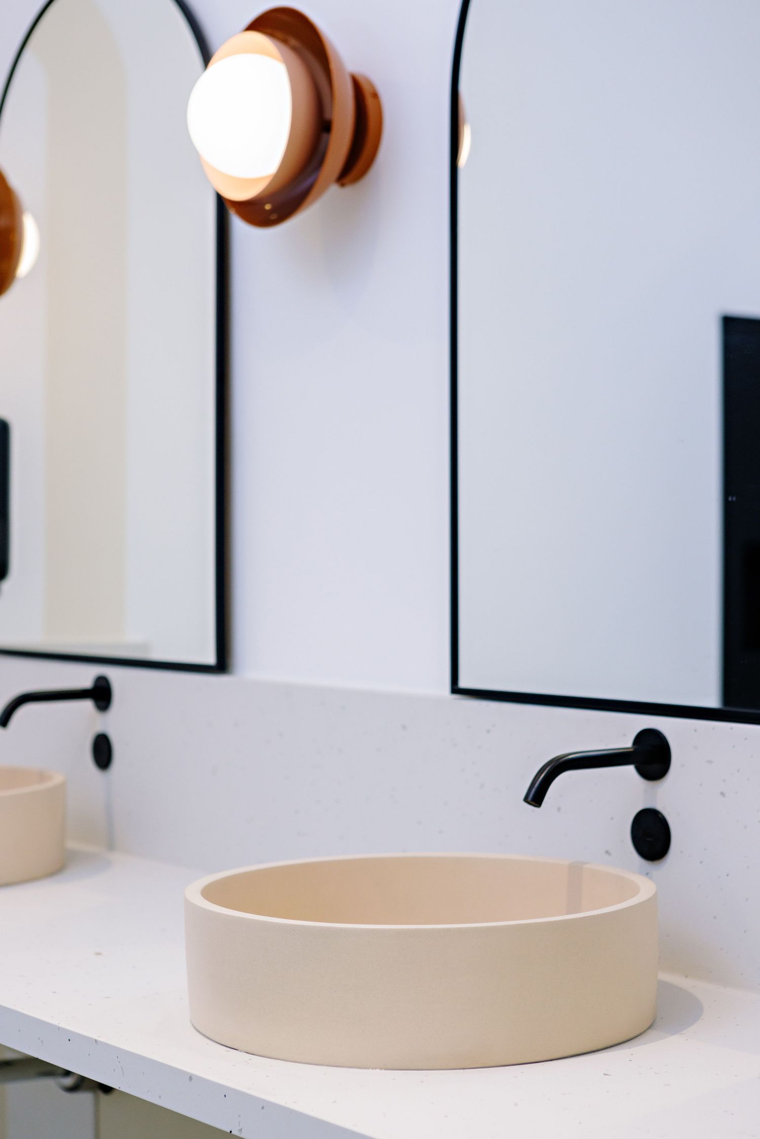 Modern bathroom interior with cream-colored vessel sink, black wall-mounted faucet, and circular wall sconce light fixture.