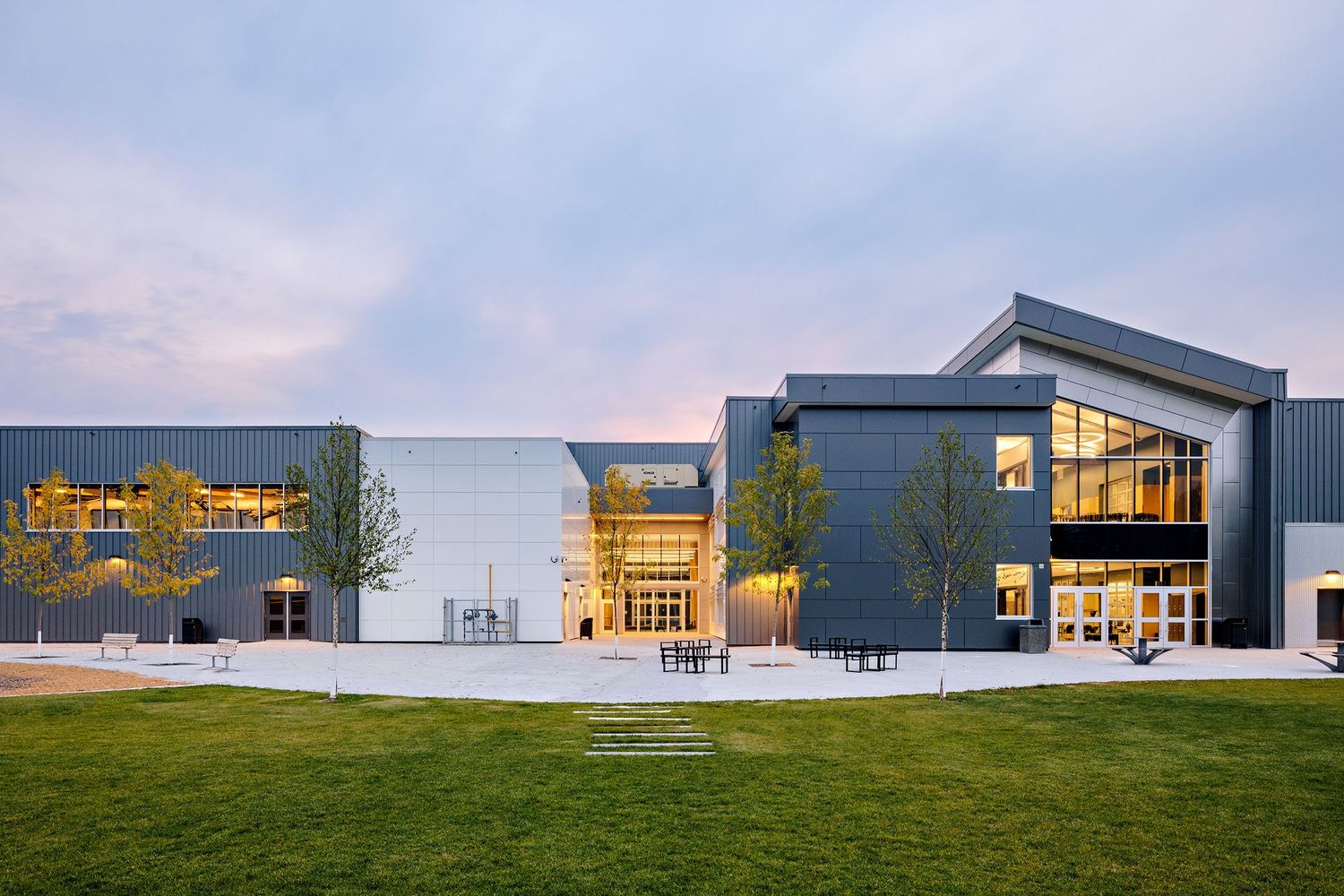 Modern school building exterior with angular architecture and glass walls illuminated at dusk against a blue sky.