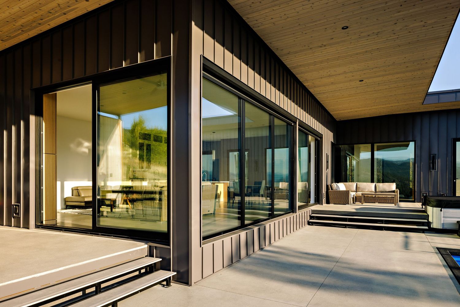 Contemporary home exterior with floor-to-ceiling windows, dark metal siding, and wraparound wooden deck under an extended roof.