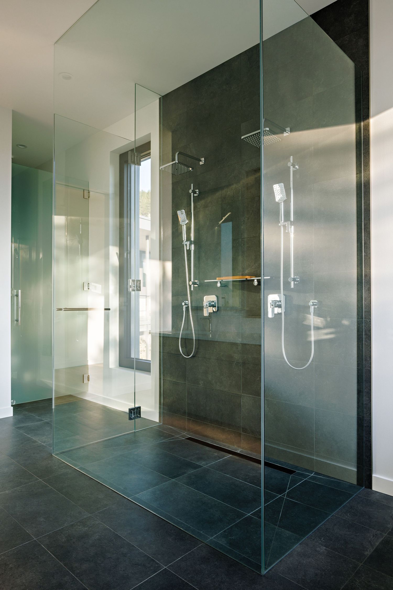 Modern glass walk-in shower with dark slate tiles, dual shower heads and floor-to-ceiling window in contemporary bathroom.
