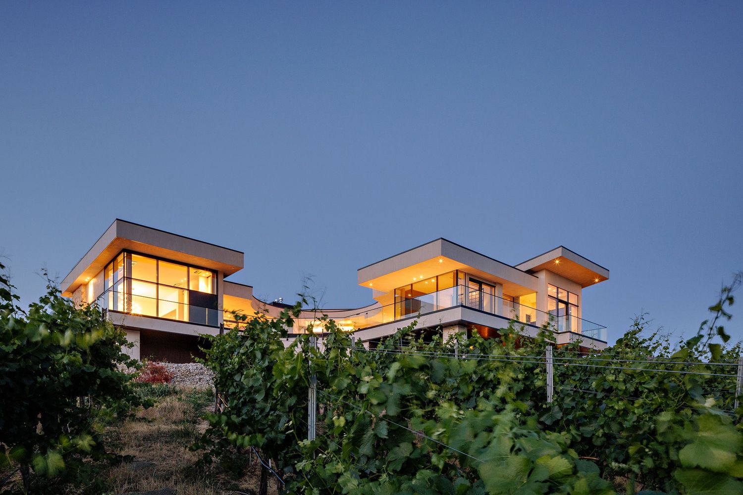 Modern hillside houses with large windows illuminated at dusk against a blue sky.