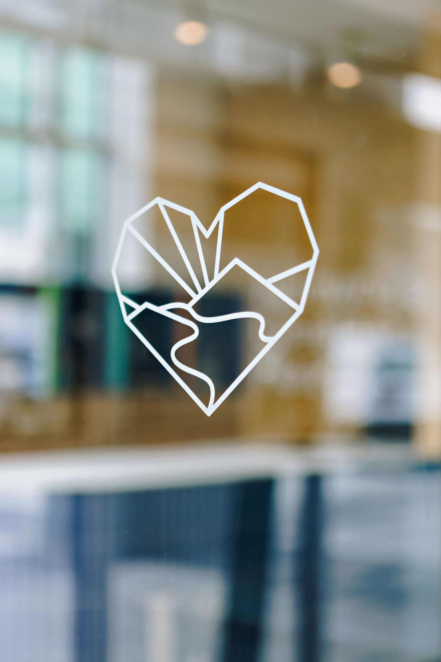 A minimalist geometric heart logo design in white lines on a blurred glass or window surface.