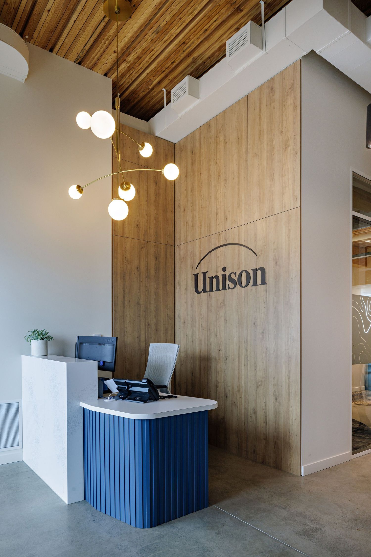 Modern office reception area with blue paneled desk, wooden wall featuring Unison logo, and contemporary globe pendant lighting.