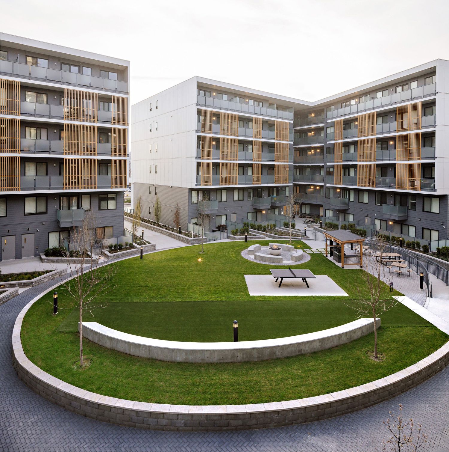 Modern apartment complex features a circular elevated green courtyard with benches and walkways surrounded by multi-story units.