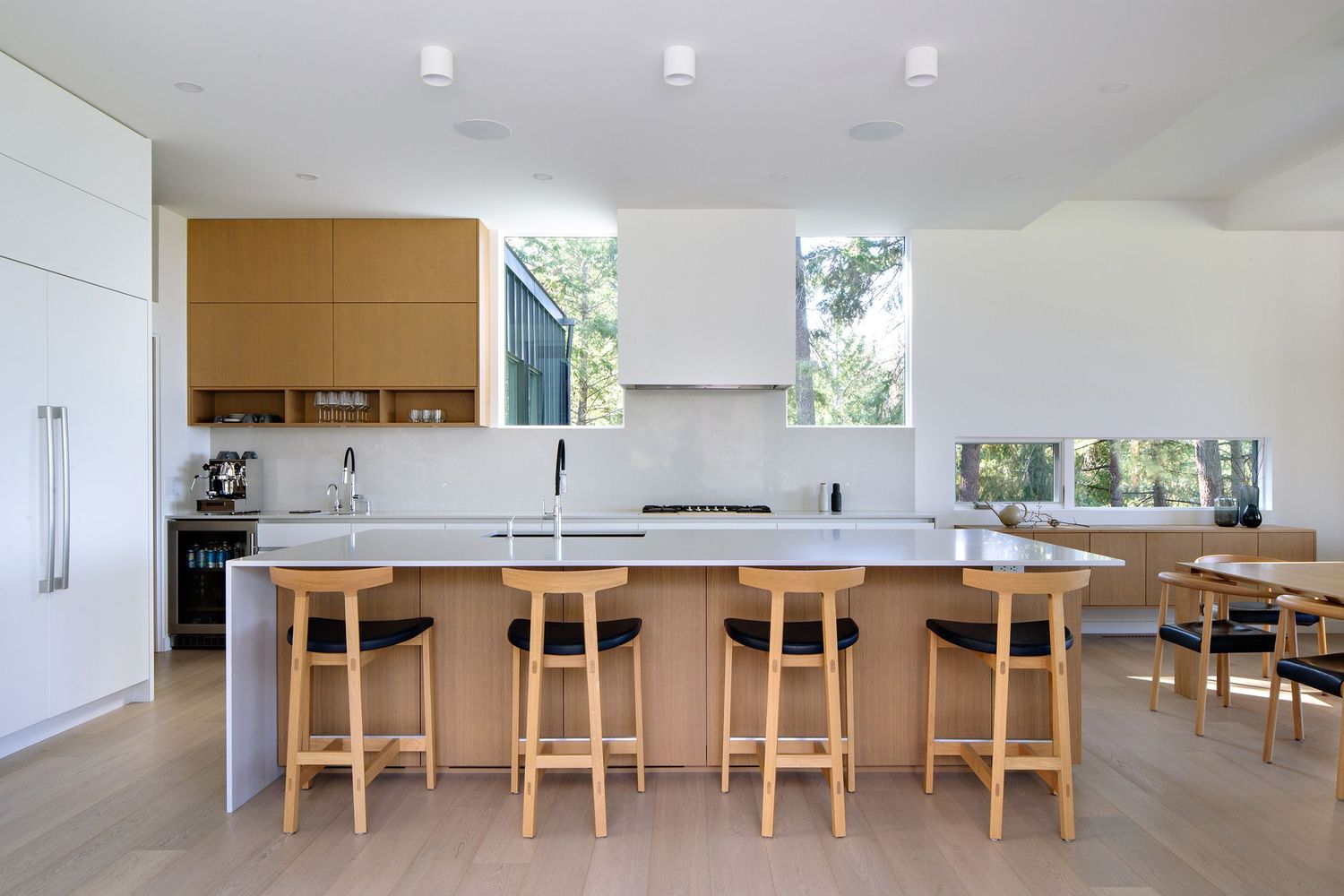 Modern open-concept kitchen with white countertops, wooden bar stools, and large windows providing natural light.