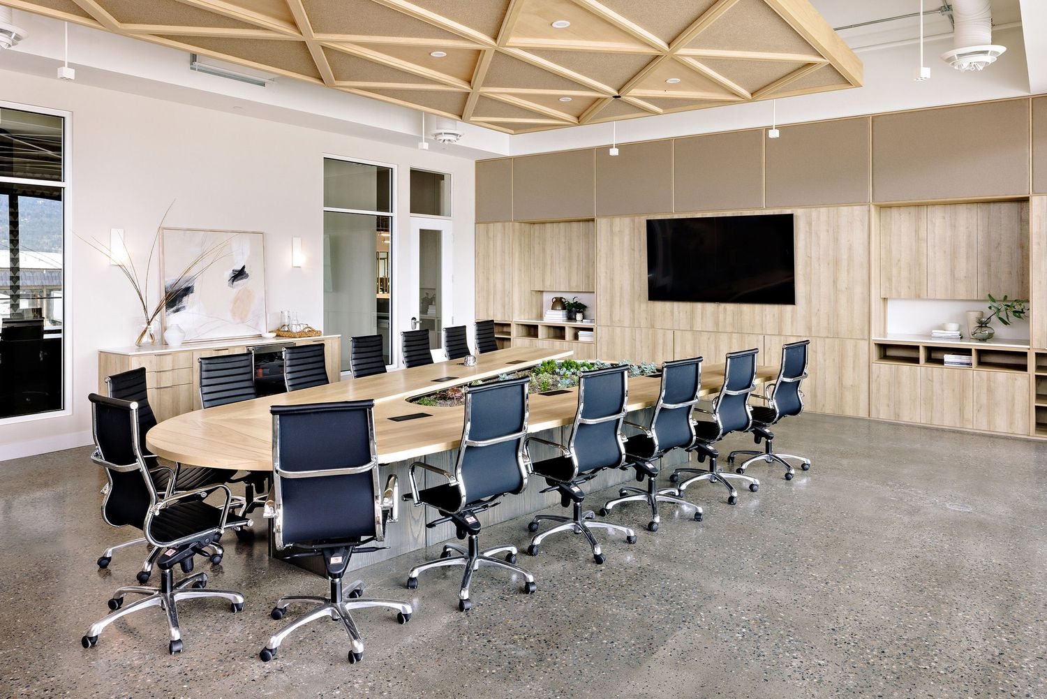 Modern conference room with large oval table, blue mesh chairs, geometric ceiling design, and wall-mounted flat screen TV.