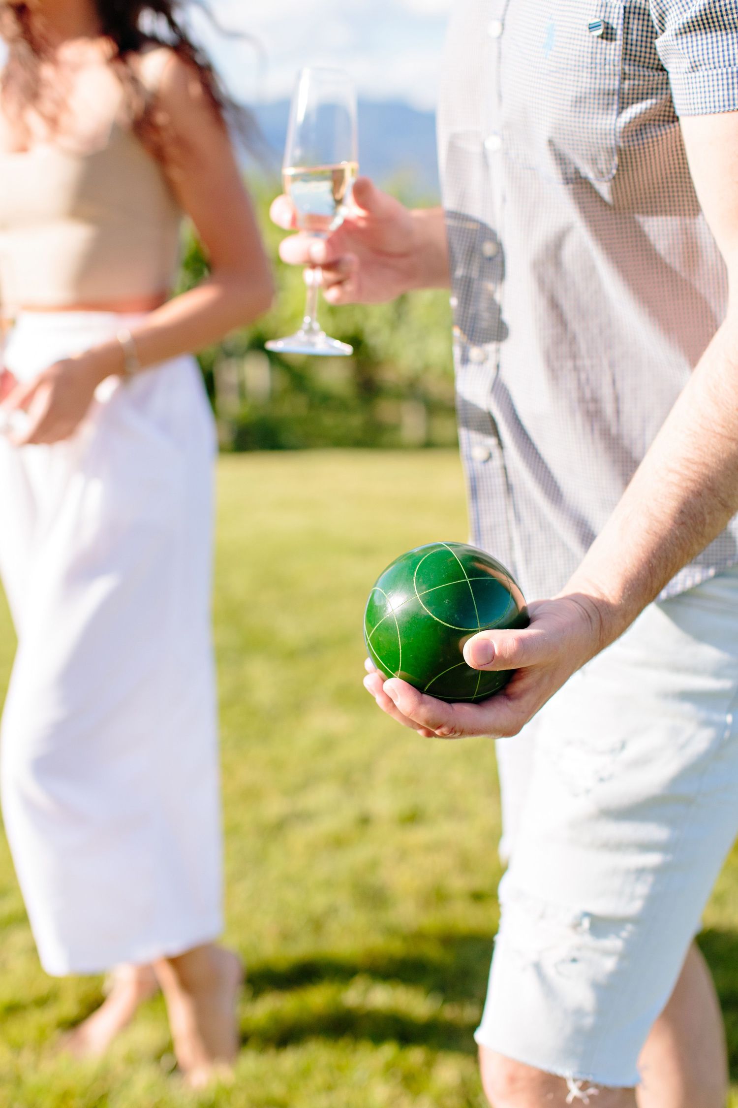 Person holding a green bocce ball and wine glass during an outdoor garden party on sunny lawn.