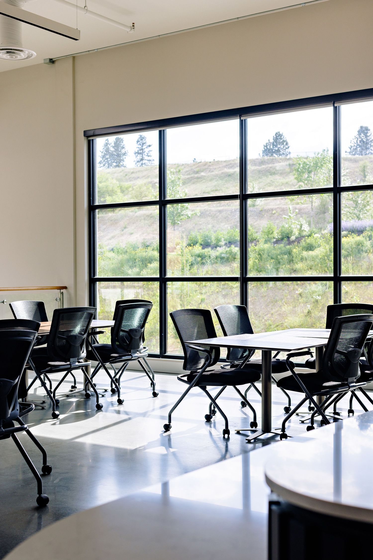 Modern classroom with black rolling chairs and white tables facing large windows overlooking a scenic hillside view.