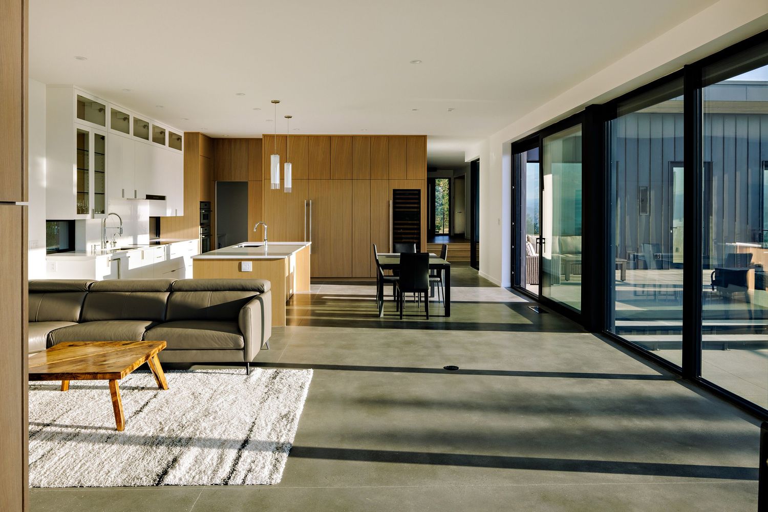 Modern open-plan living space with wood paneling, floor-to-ceiling windows, and minimalist furniture on a geometric patterned rug.