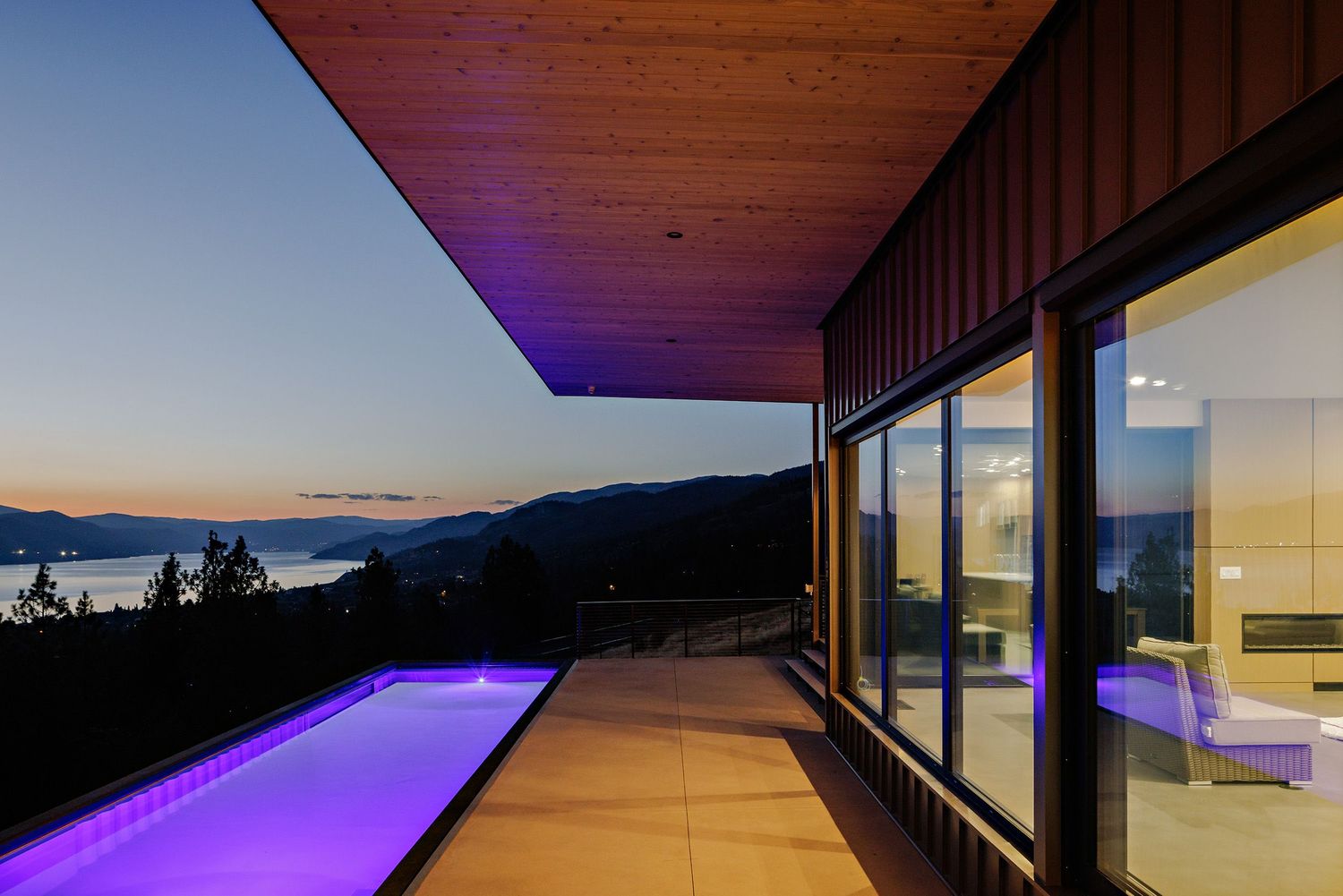 Modern mountain home at dusk with purple LED accent lighting illuminating a covered deck and large glass windows.