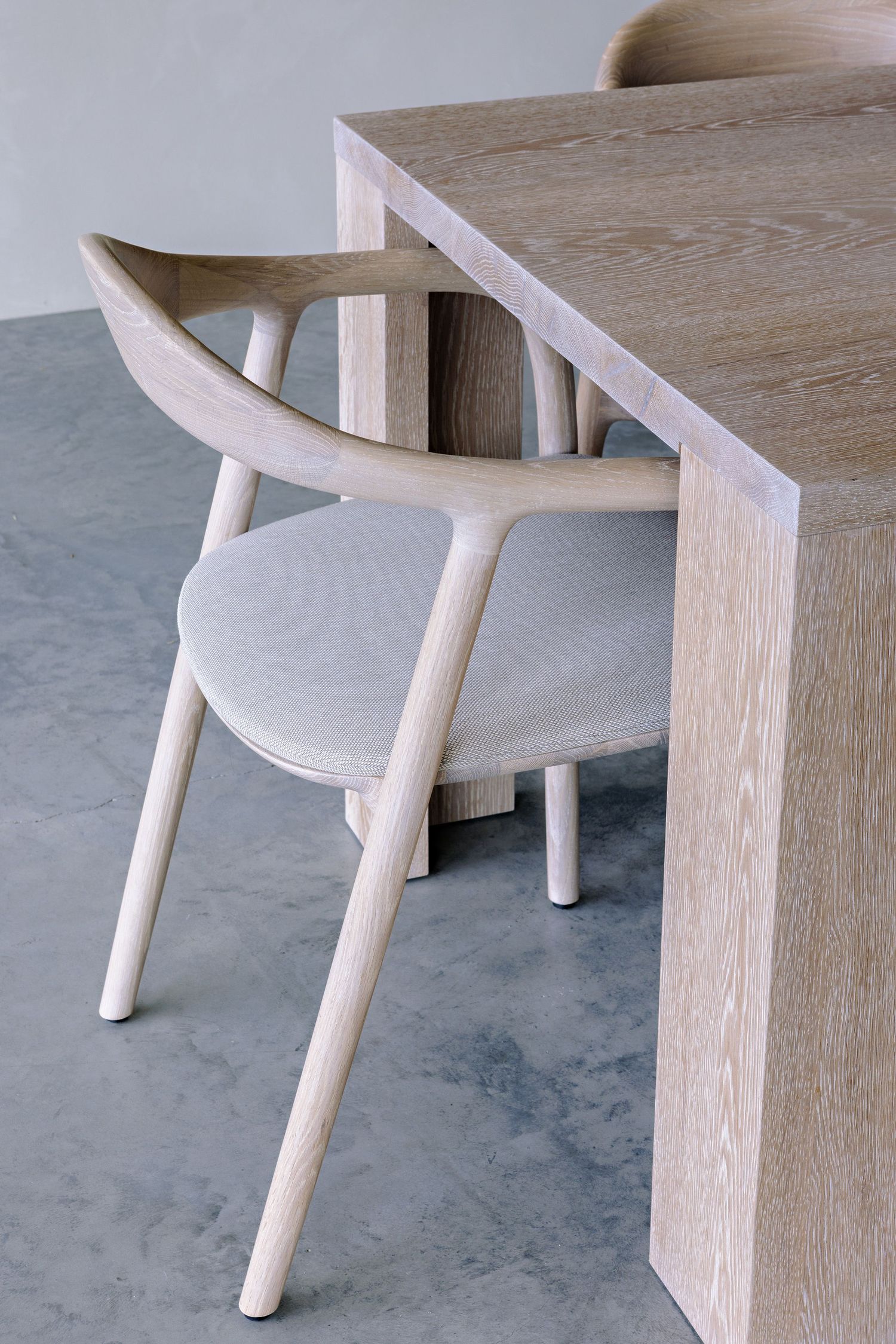 Modern minimalist wooden dining chair with curved backrest and angled armrests shown against light wood table on concrete floor.