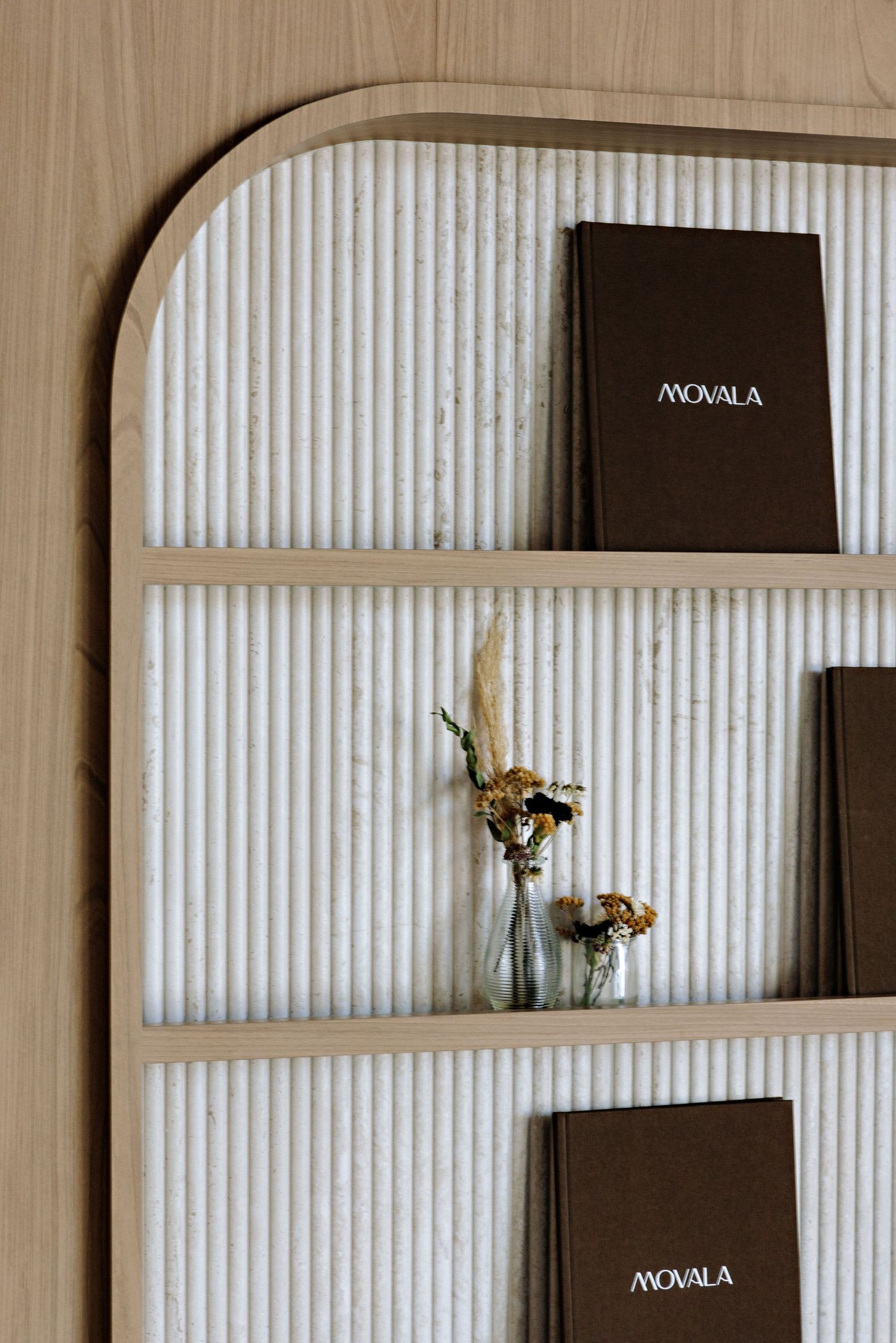 Minimalist wooden shelf with fluted textured panel displays dark brown Akoala books and dried flowers in glass vase.