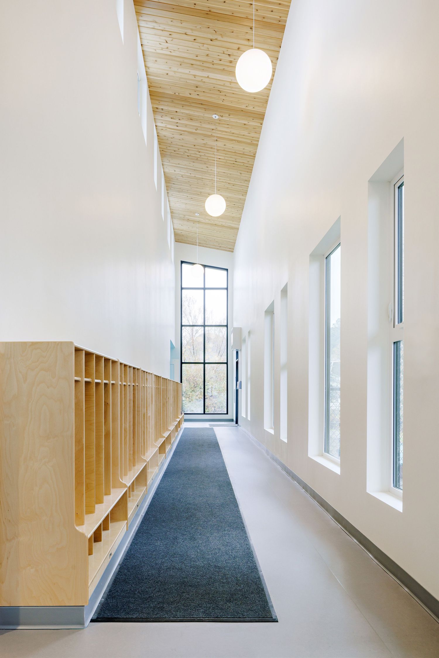Long corridor with sloped wooden ceiling, white walls, blue carpet and natural light streaming through windows.