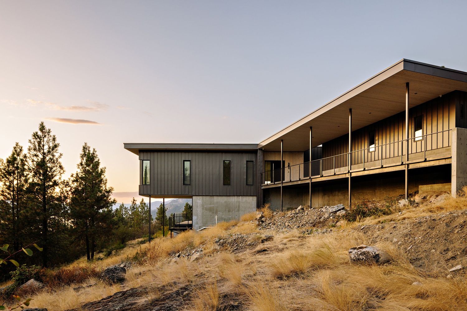 Modern hillside home with wooden deck and large windows overlooks mountain landscape at sunset.