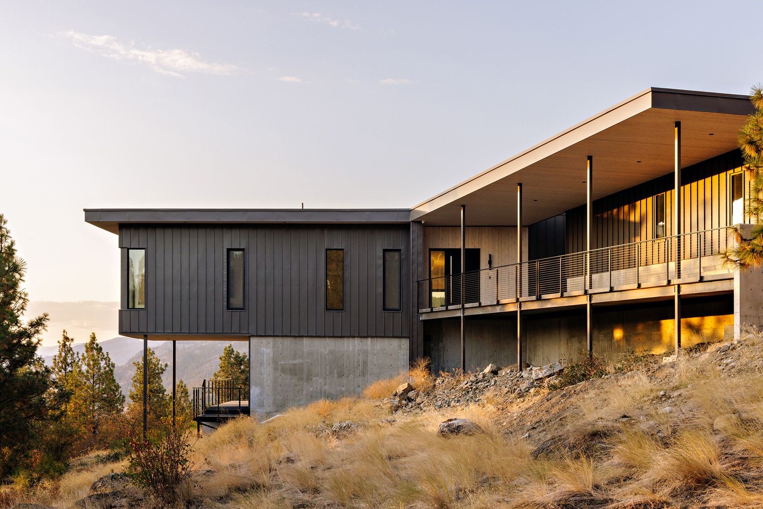 Modern hillside home with gray metal siding and extended deck overlooking scenic mountain landscape at sunset.