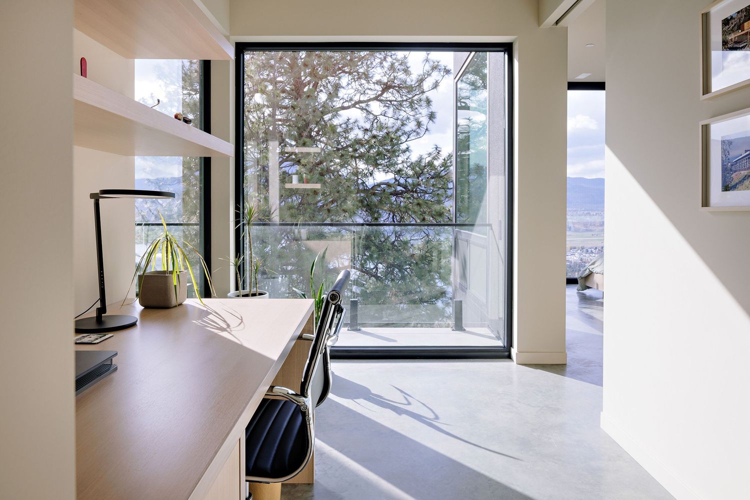 Modern home office workspace with large window overlooking trees, featuring built-in desk and natural lighting.