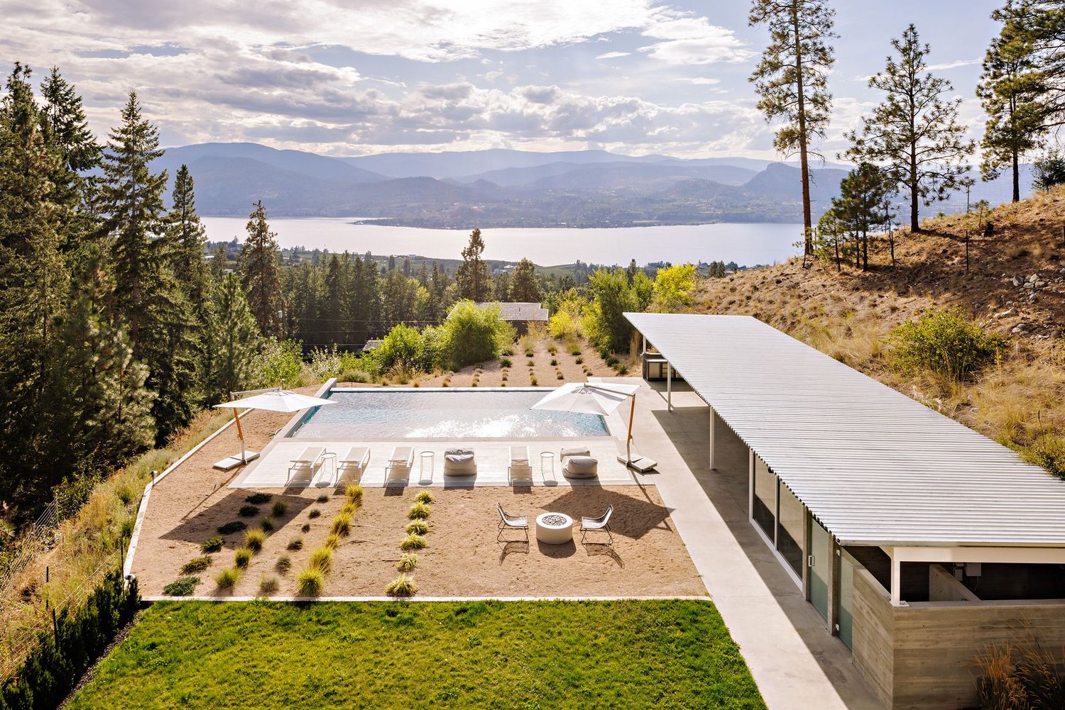 Modern hillside home with panoramic lake views features a sleek patio and geometric landscaping design overlooking the forest.