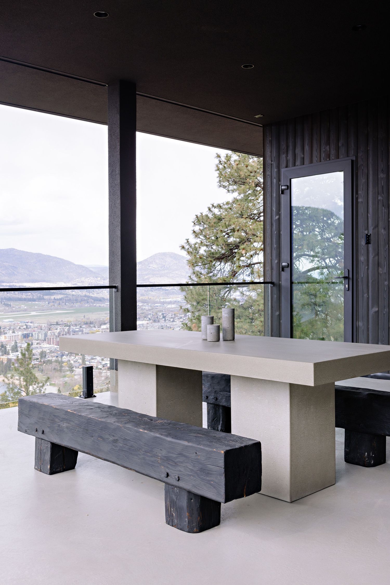 Modern concrete dining table with rustic wooden benches on covered patio overlooking mountain valley landscape.