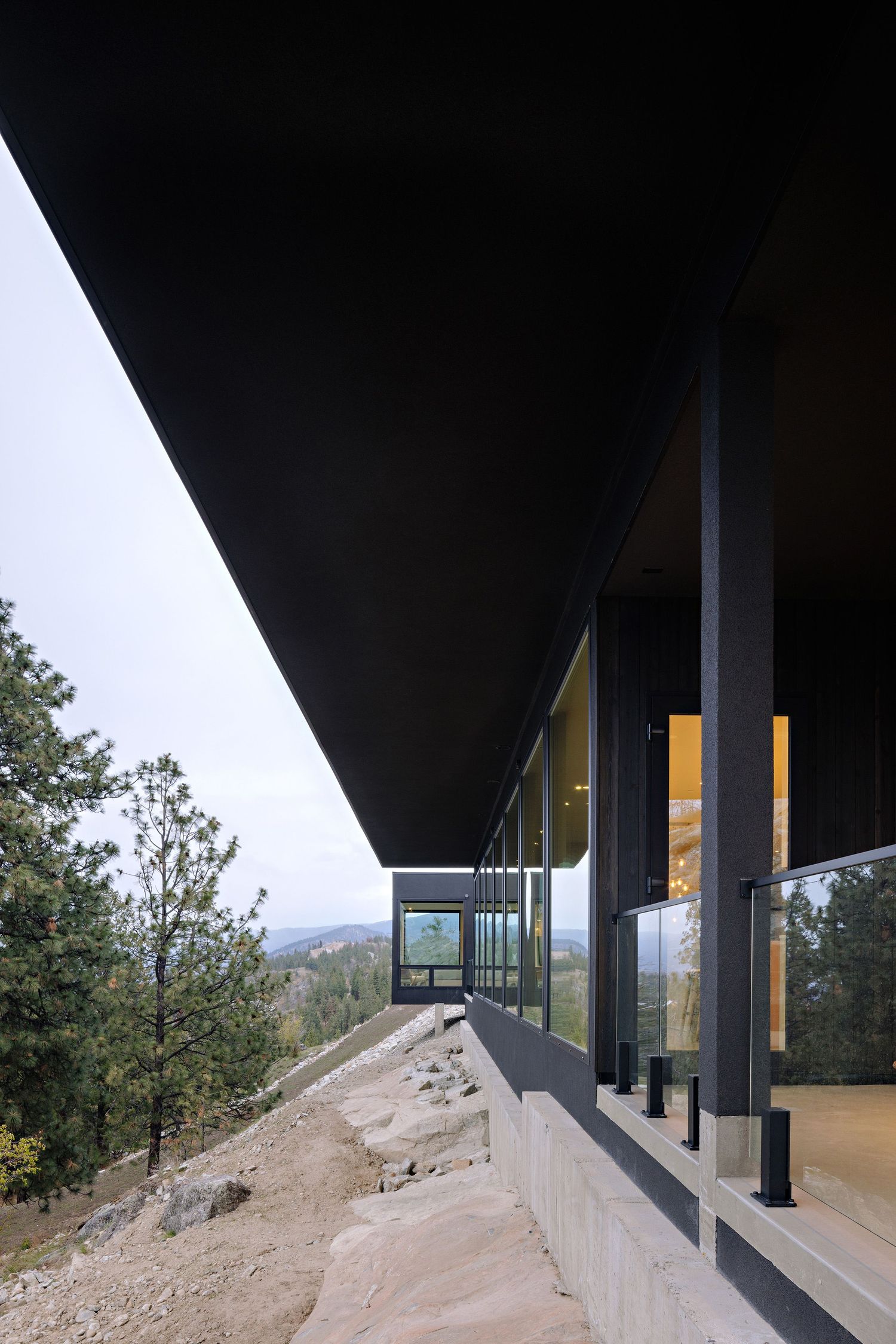 Modern architectural building with floor-to-ceiling windows and dramatic black overhang positioned on a rocky mountain slope.