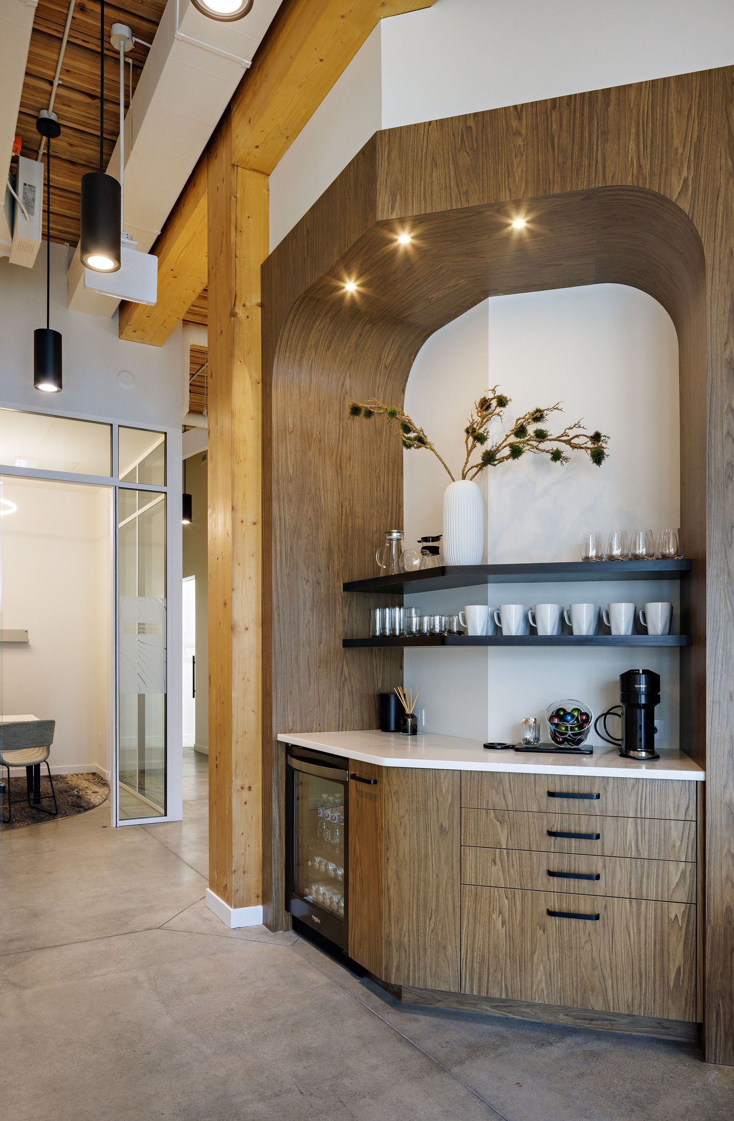 Modern office kitchenette with curved wood paneling, built-in shelving, and exposed wooden beam ceiling details.