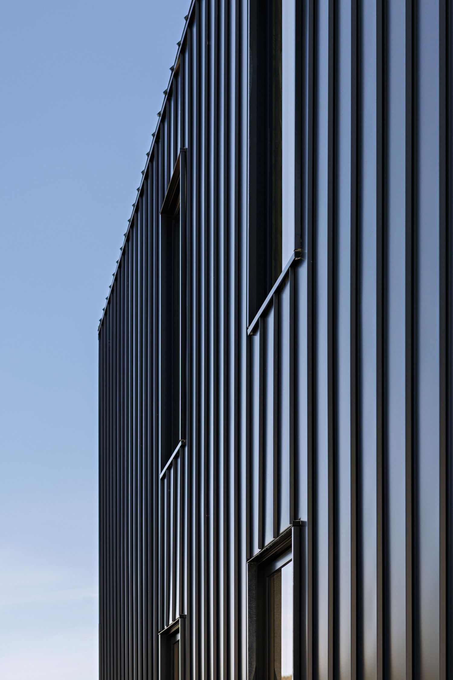 Modern industrial building facade with vertical black metal panels and diagonal light fixtures against blue sky.