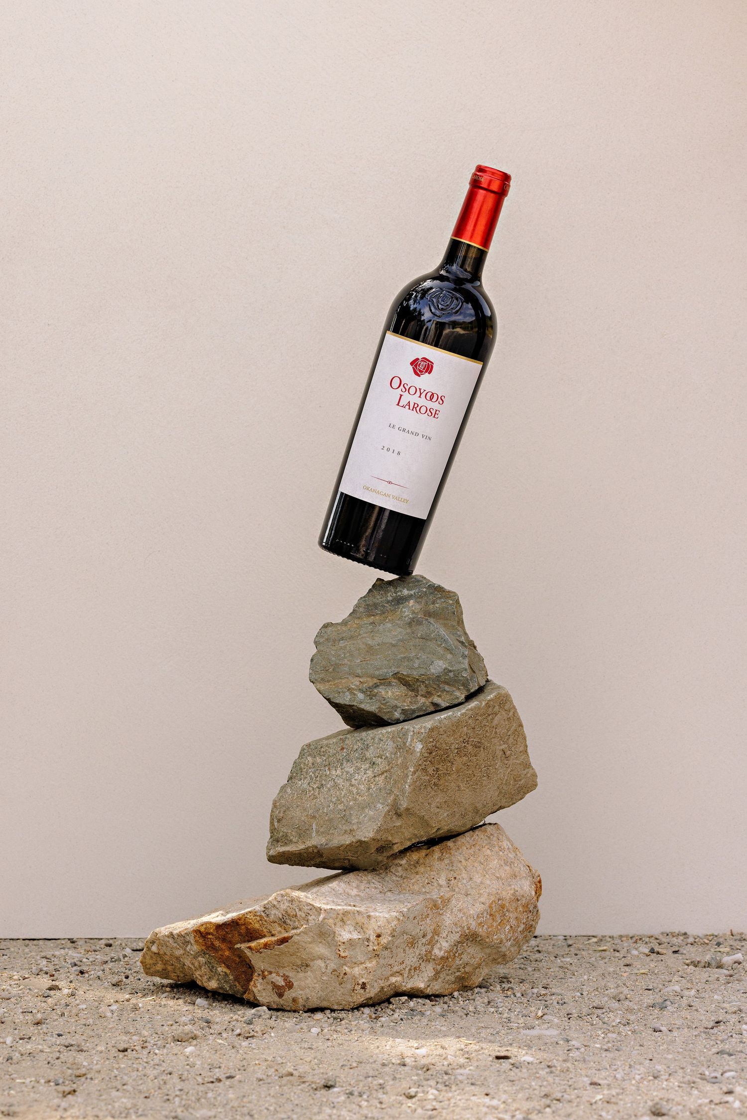 A red wine bottle precariously balanced on a stack of rough stones against a beige background.