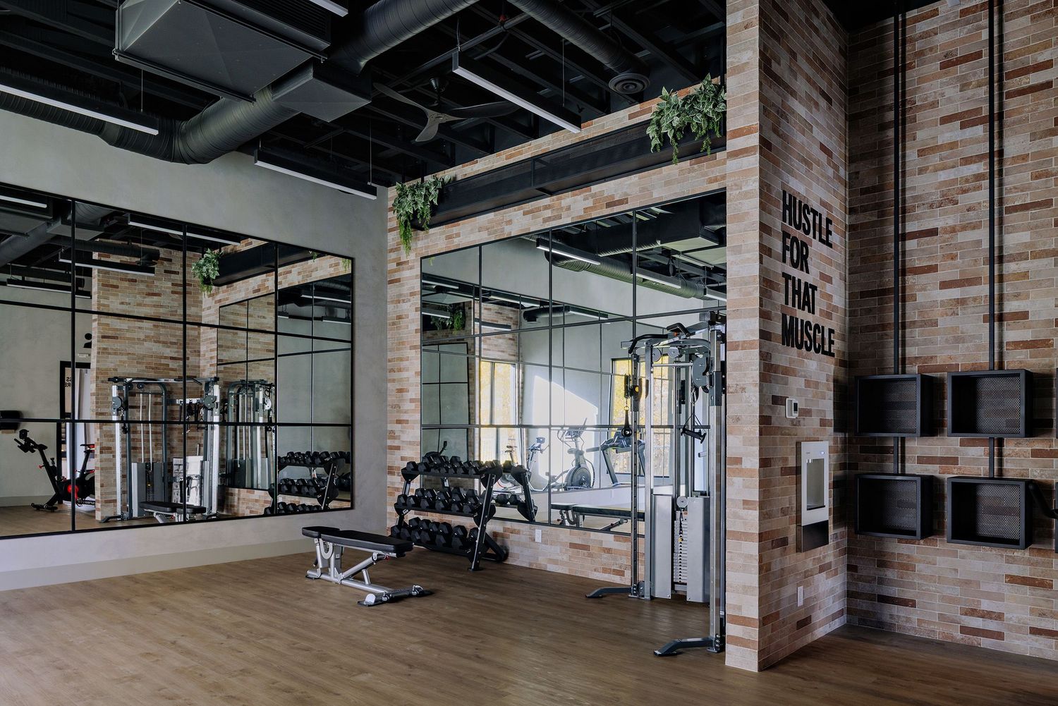 Modern industrial-style gym interior with exposed brick walls, high ceilings, and fitness equipment arranged throughout the space.