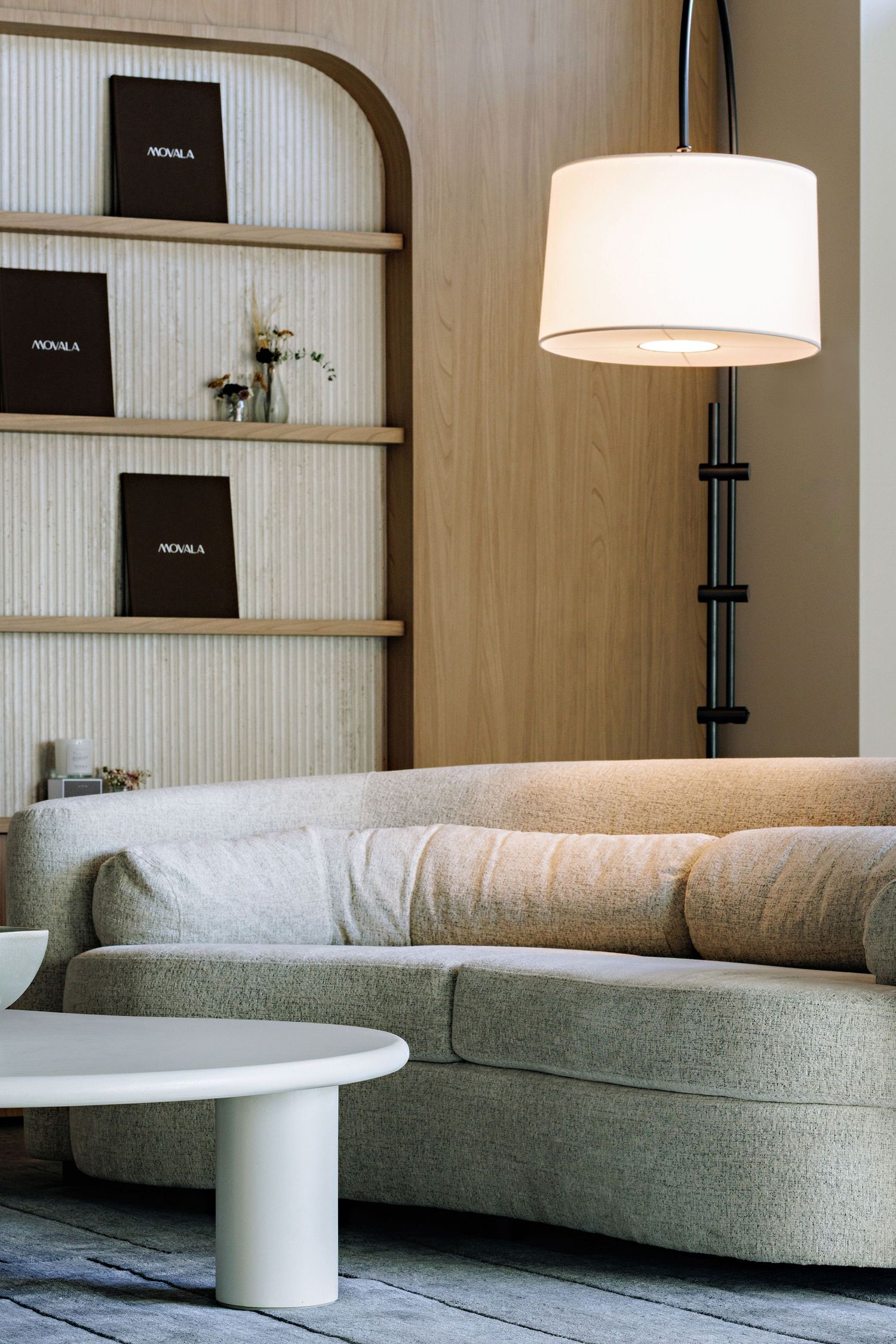 Modern living room with curved sofa, white lamp, and built-in shelving displaying black SENSA books on wooden wall.