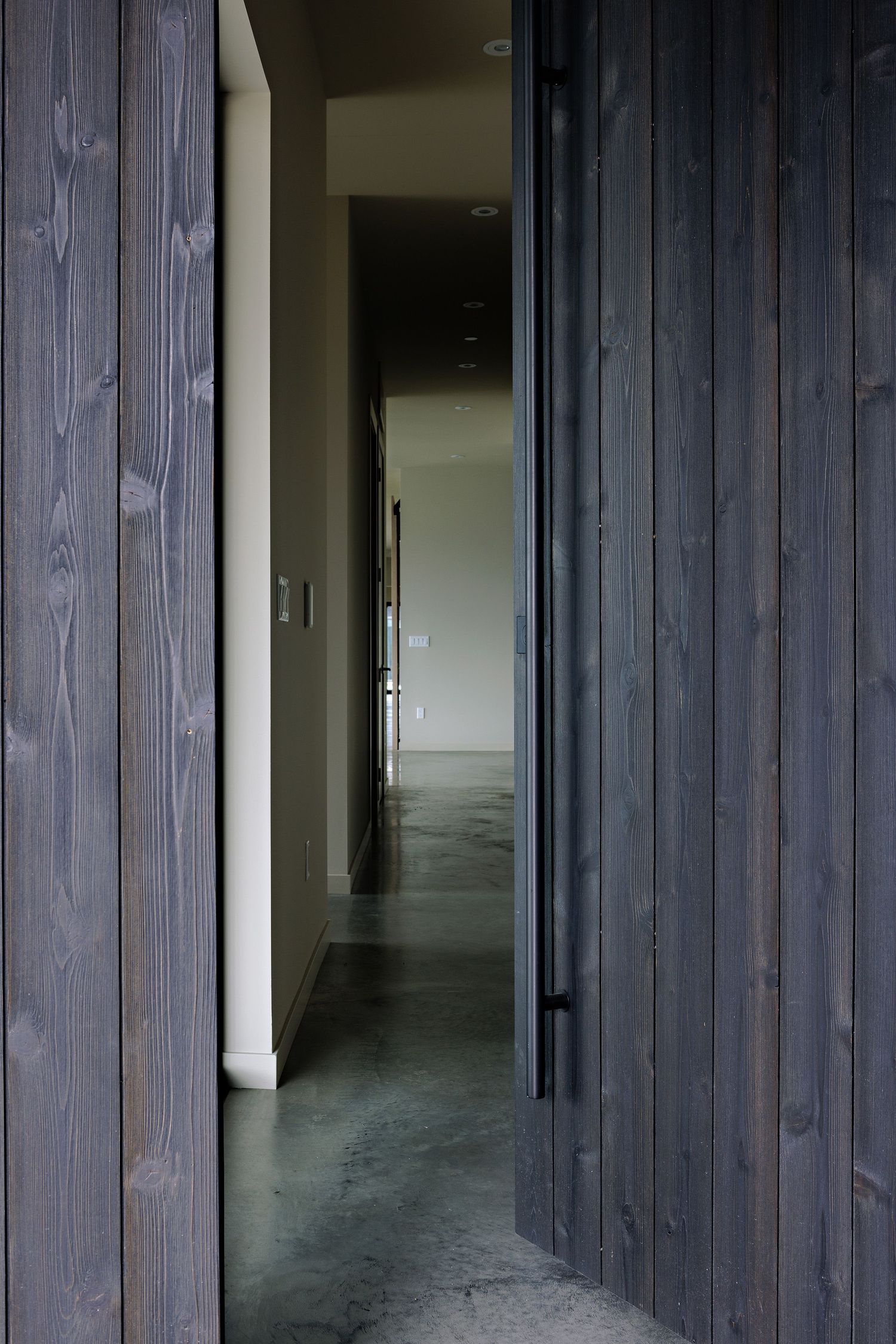 Dark wood-paneled hallway with concrete floors leads into a minimalist modern interior space.