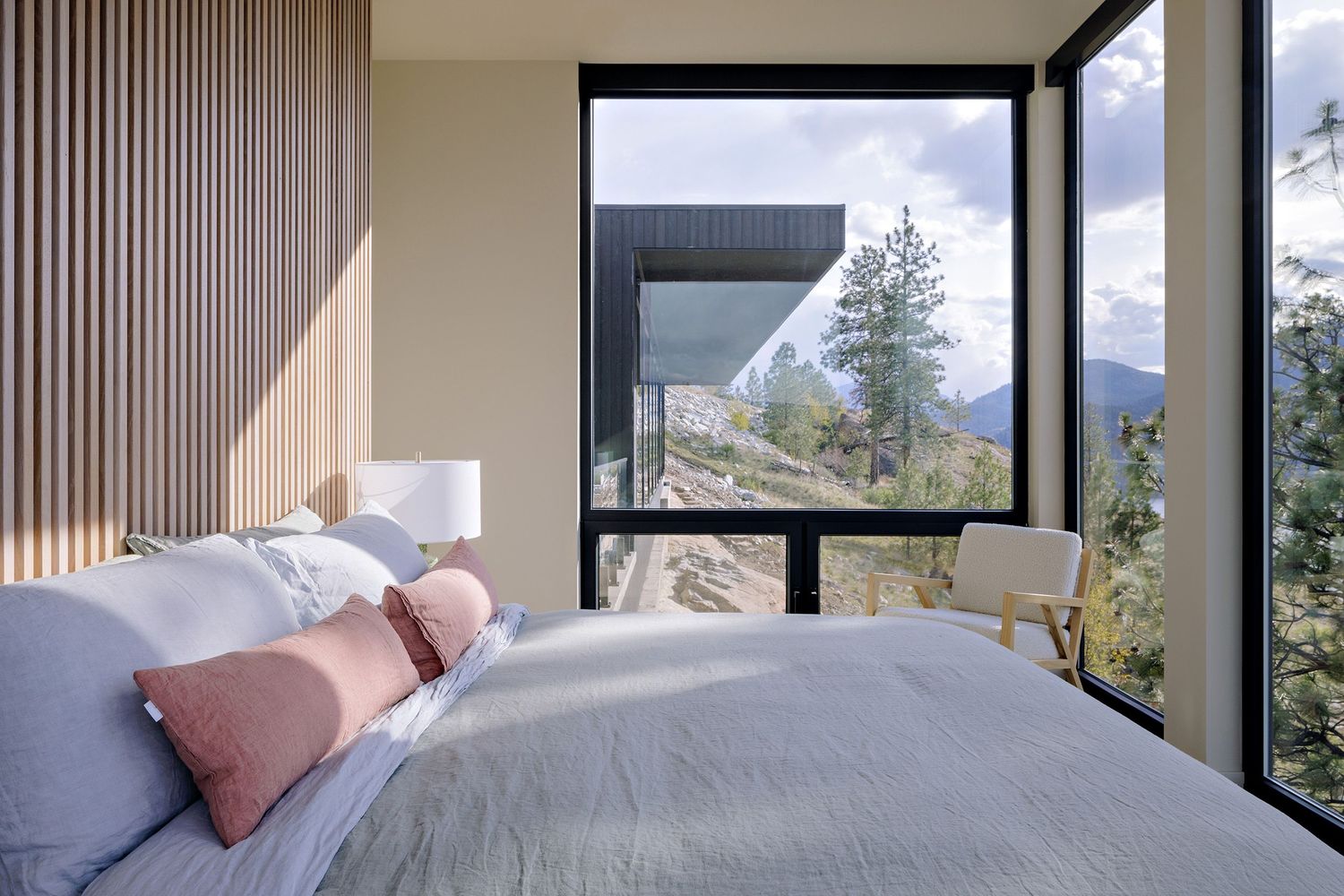 Modern bedroom with floor-to-ceiling windows offering scenic mountain views and gray bedding with pink accent pillows.