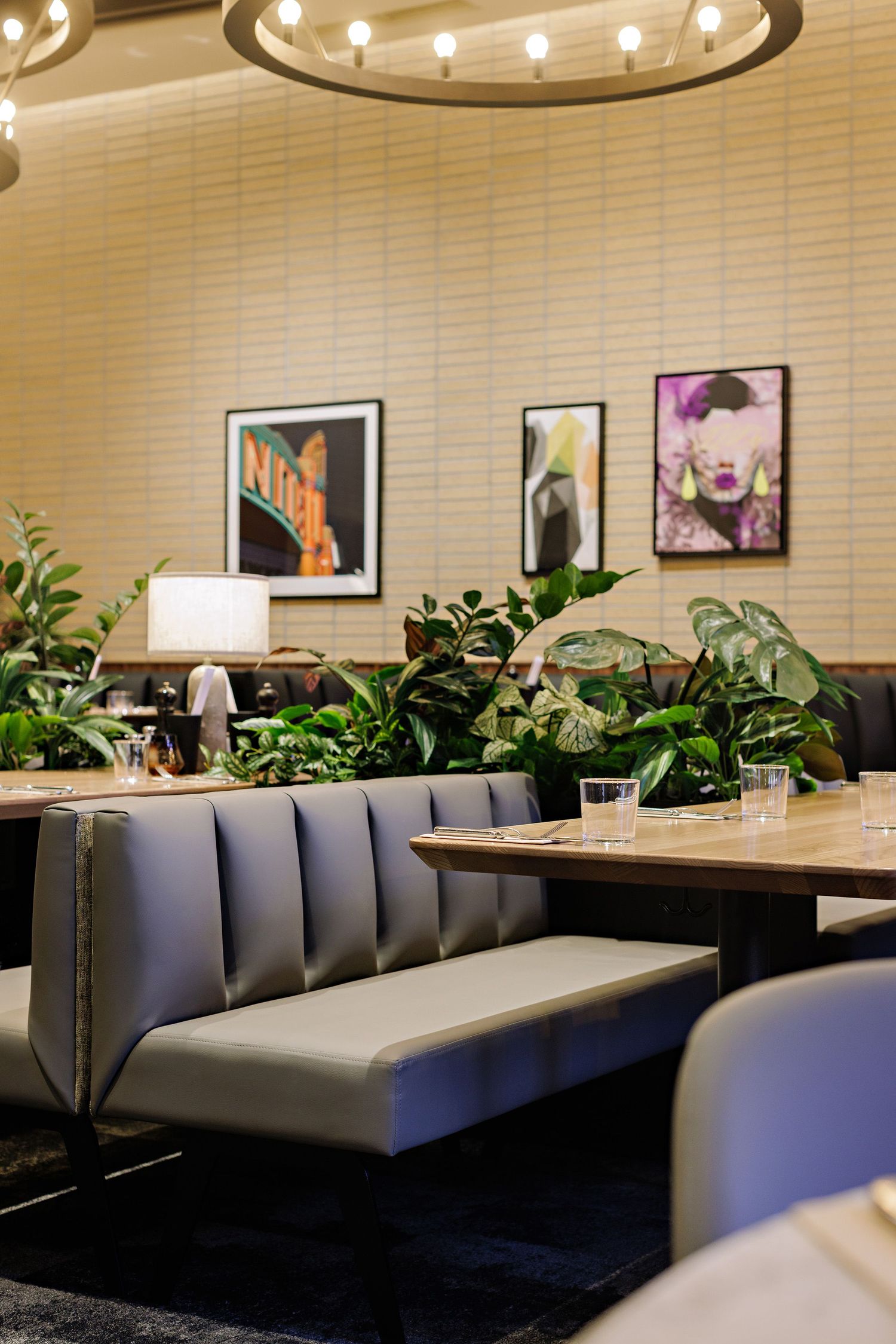 Modern restaurant interior with grey booth seating, potted plants, and framed artwork on cream-colored walls under circular lighting.