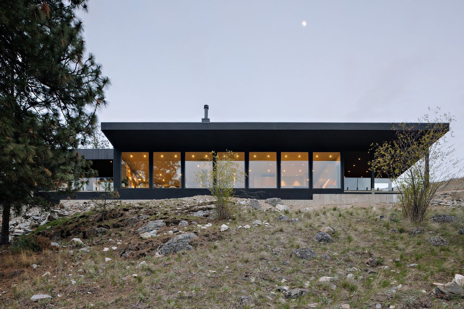 Modern hillside house with large glass windows and black exterior facade perched on rocky terrain at dusk.