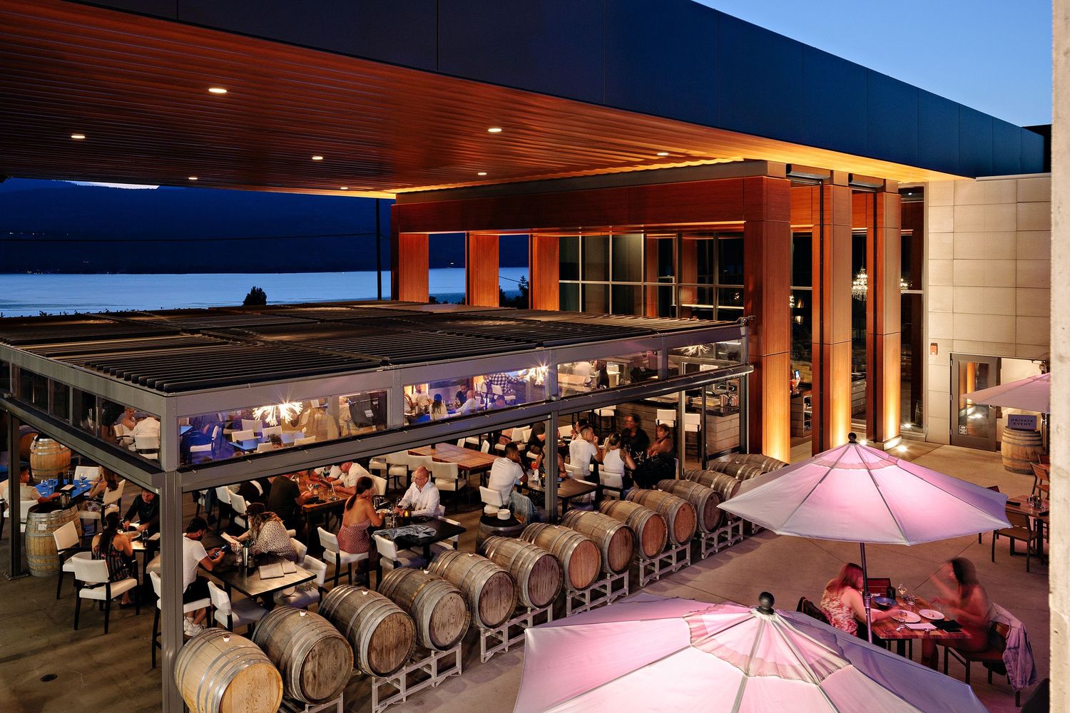 Outdoor restaurant patio with wood ceiling features illuminated bar seating and pink umbrellas at sunset.