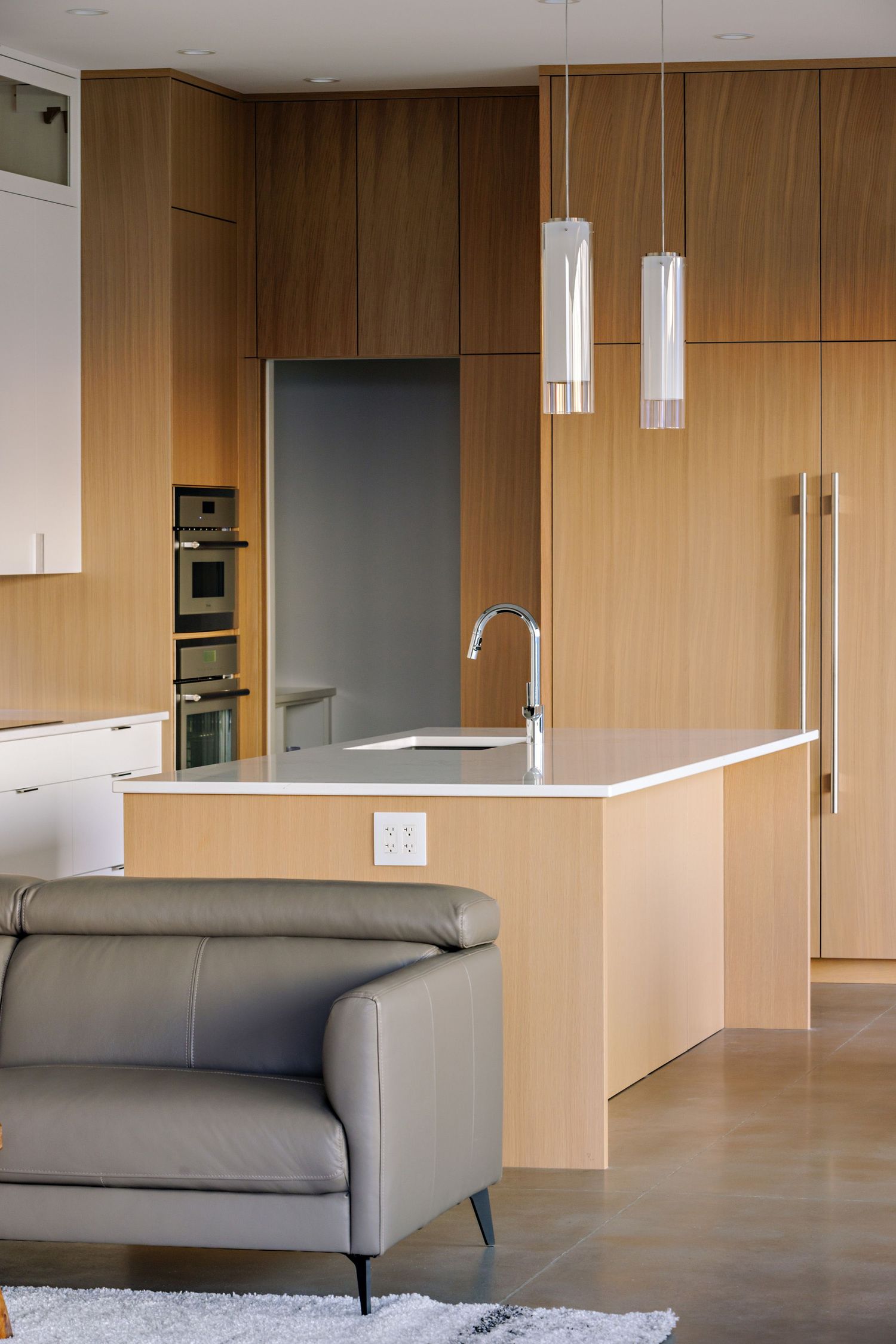 Modern kitchen features light wood cabinetry, pendant lights, and a gray sofa in an open-concept living space.