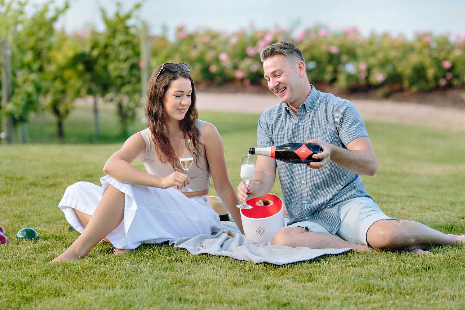 A couple enjoys a sunny picnic on a blanket in a garden with fresh fruit and drinks.